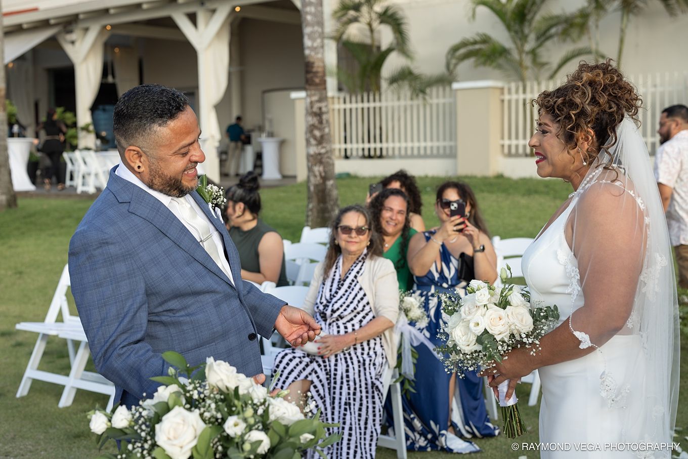 Couple exchanges vows at outdoor wedding ceremony, bride holding white floral bouquet, guests watching in background.