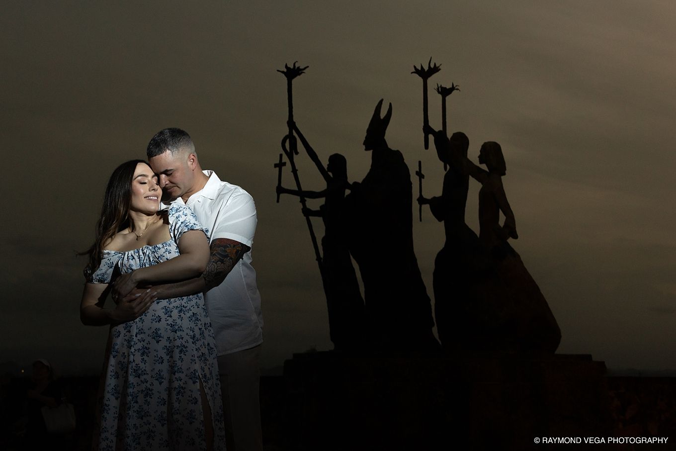 Couple embracing at dusk with dramatic silhouette statues of robed figures holding staffs in the background. - Love Story