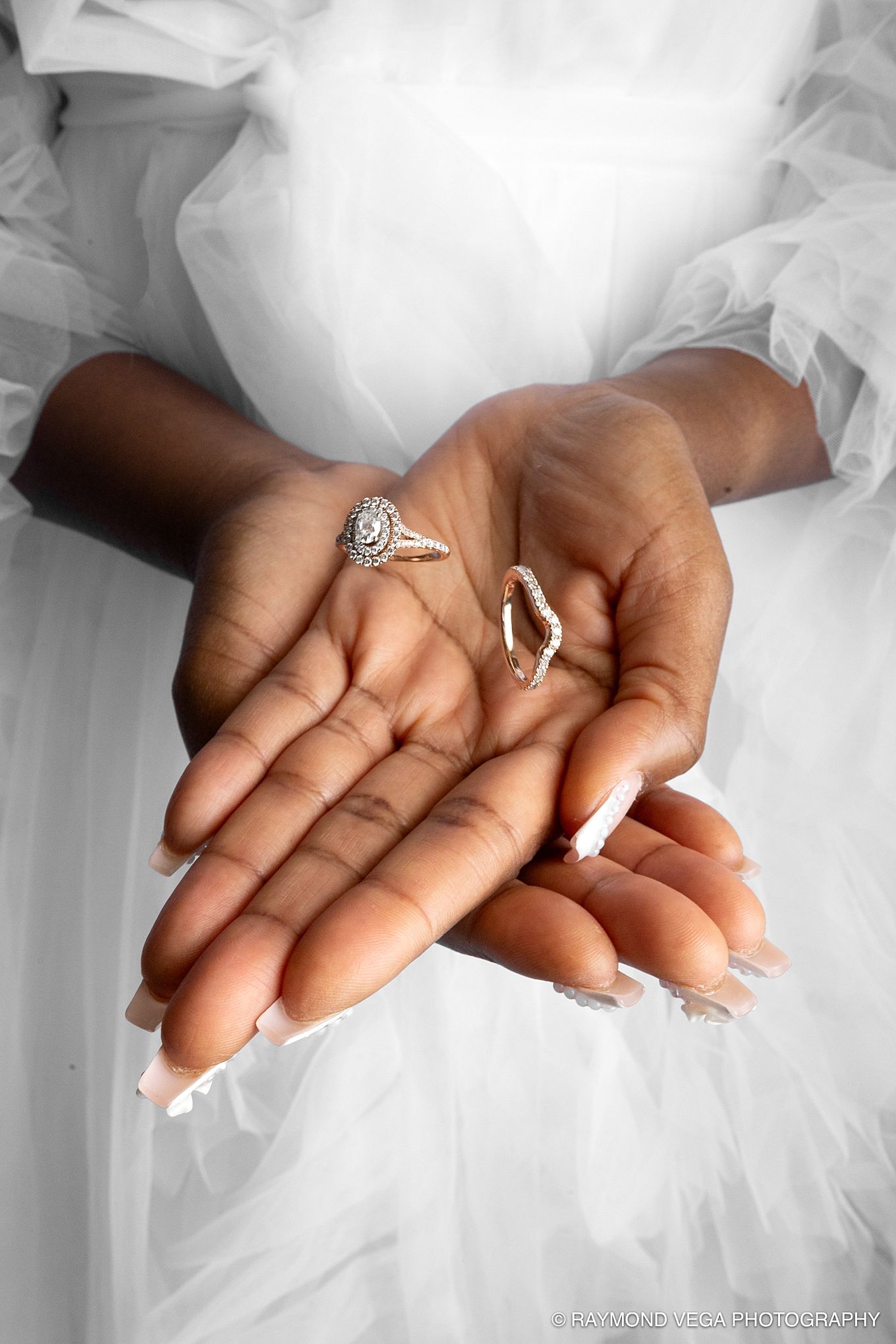 Bride with French manicure holds diamond engagement ring and wedding band in cupped hands against white bridal gown. Hotel Condado Vanderbil