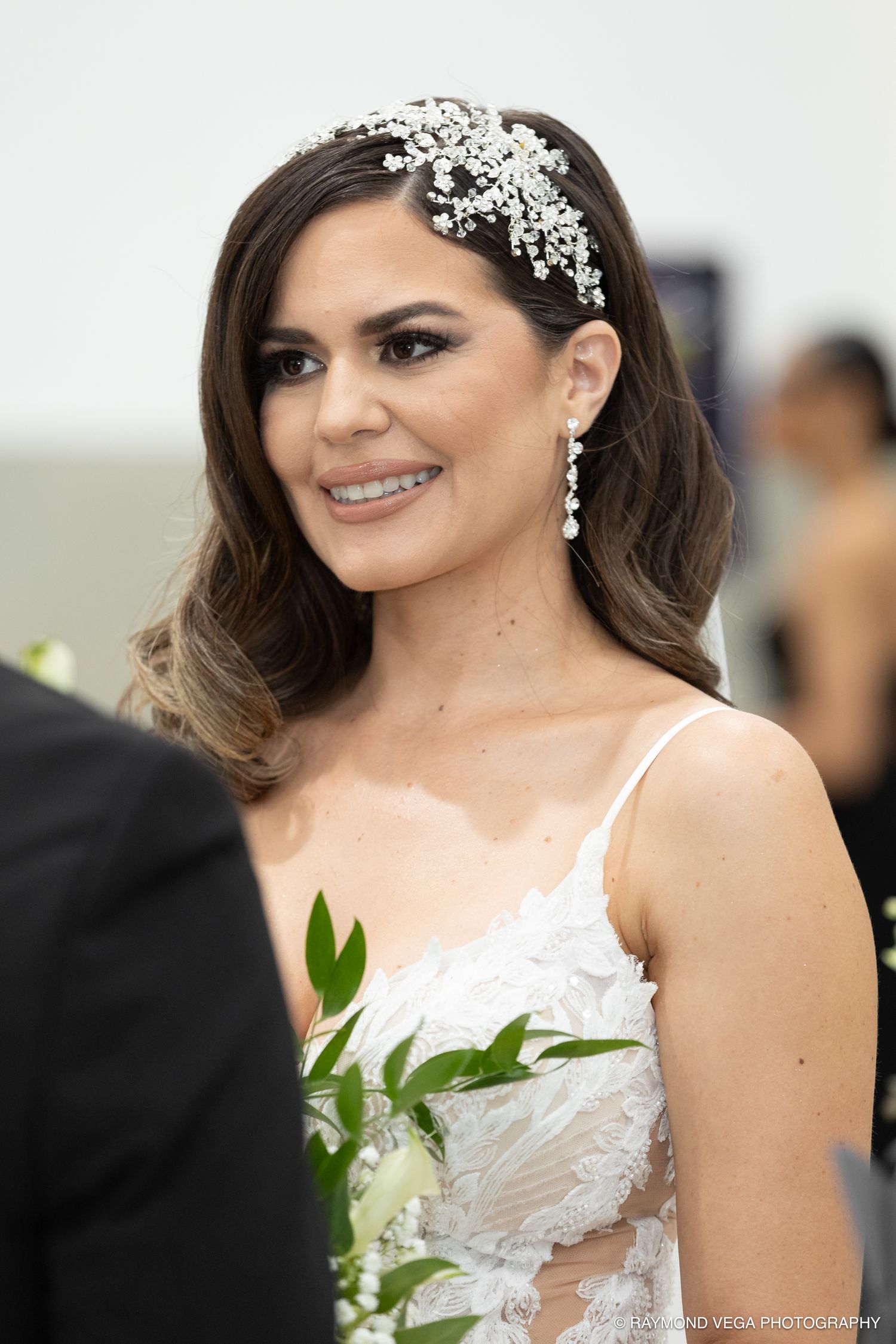 Smiling bride in white lace gown wearing crystal hair piece and drop earrings holding white flowers at wedding ceremony.