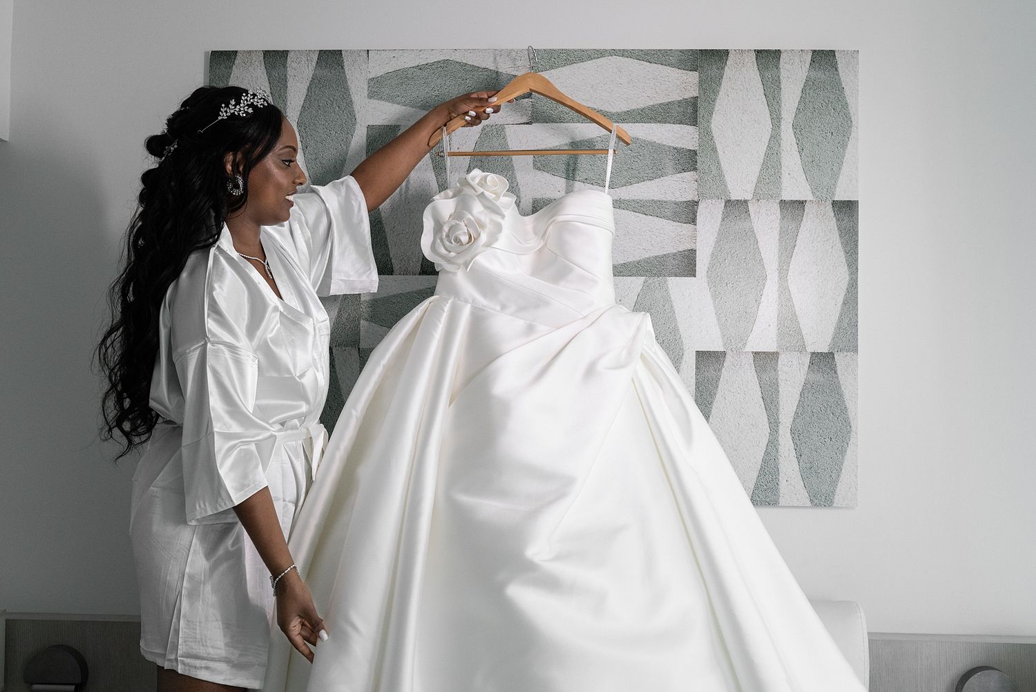 Bride in white robe admiring her elegant ball gown wedding dress on a hanger in a bright bridal suite.