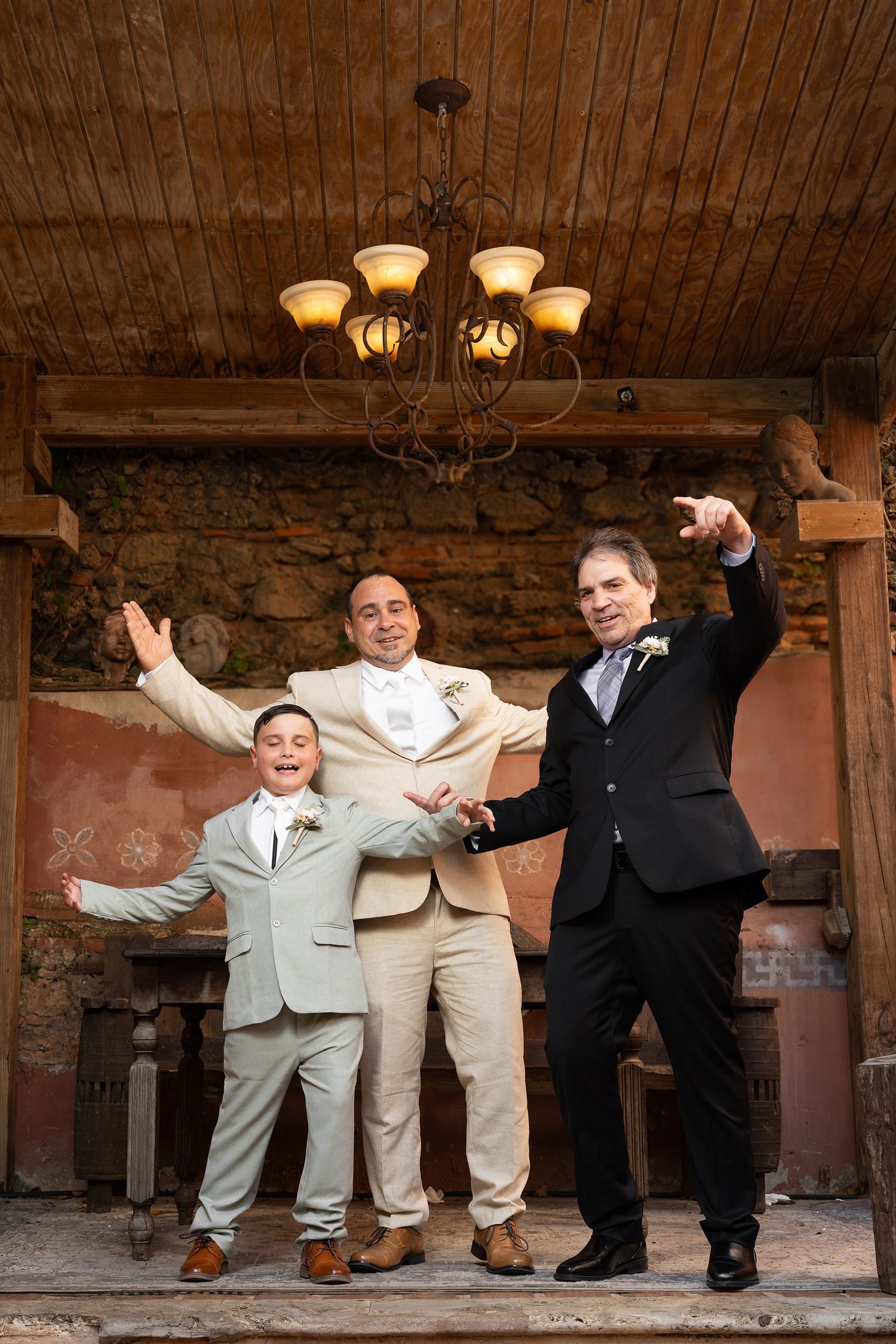 Two grooms and a young boy in formal suits celebrate joyfully inside a rustic stone venue with chandelier.