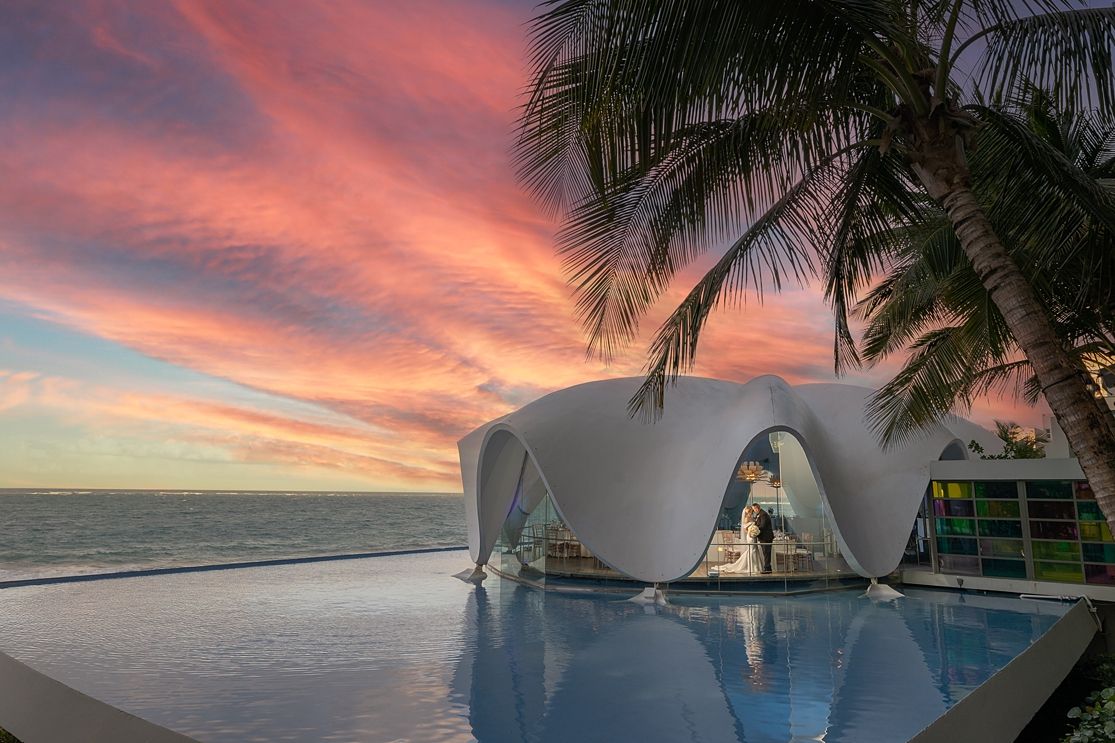 Stunning beachfront infinity pool at sunset with a modern white dome structure and palm trees against a vibrant pink sky. Hotel La Concha