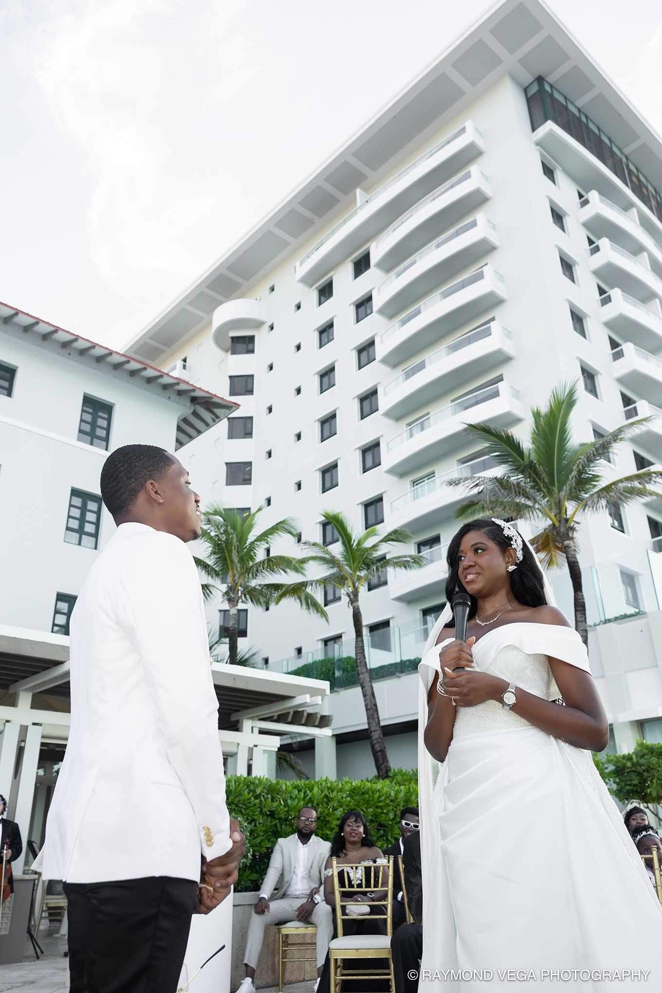 Bride and groom exchange vows outdoors at a tropical hotel wedding venue with palm trees and white building backdrop.