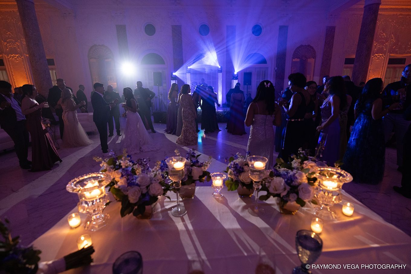 Elegant wedding reception with purple uplighting, candlelit floral centerpieces, and guests dancing on a grand ballroom floor.Antiguo Casino