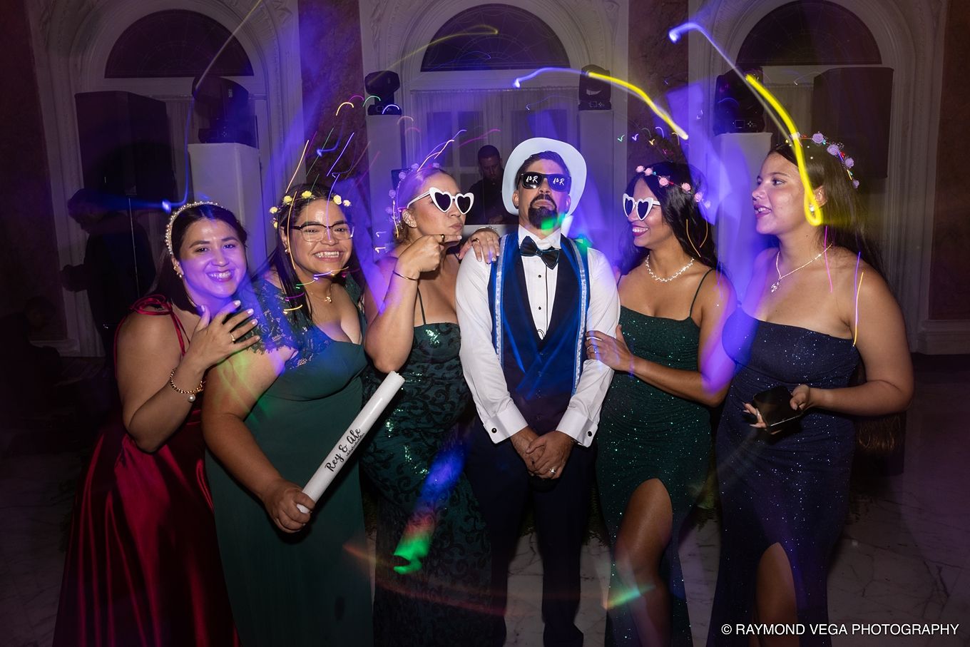 Group of five friends posing at a prom or formal event, wearing elegant gowns and fun glasses with glowing accessories. Antiguo Casino
