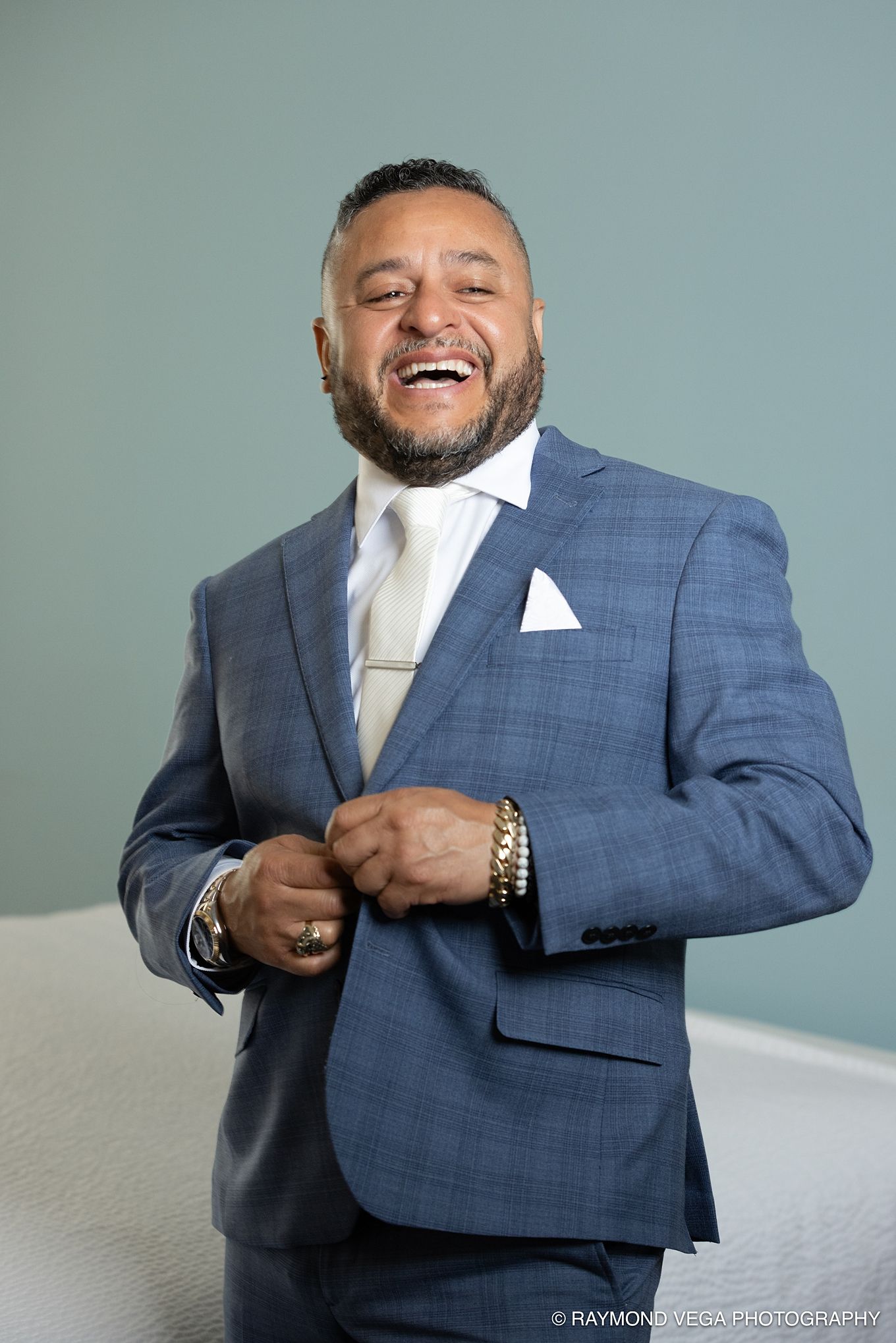Laughing man in blue plaid suit with white tie and pocket square, adjusting jacket buttons at a formal event.