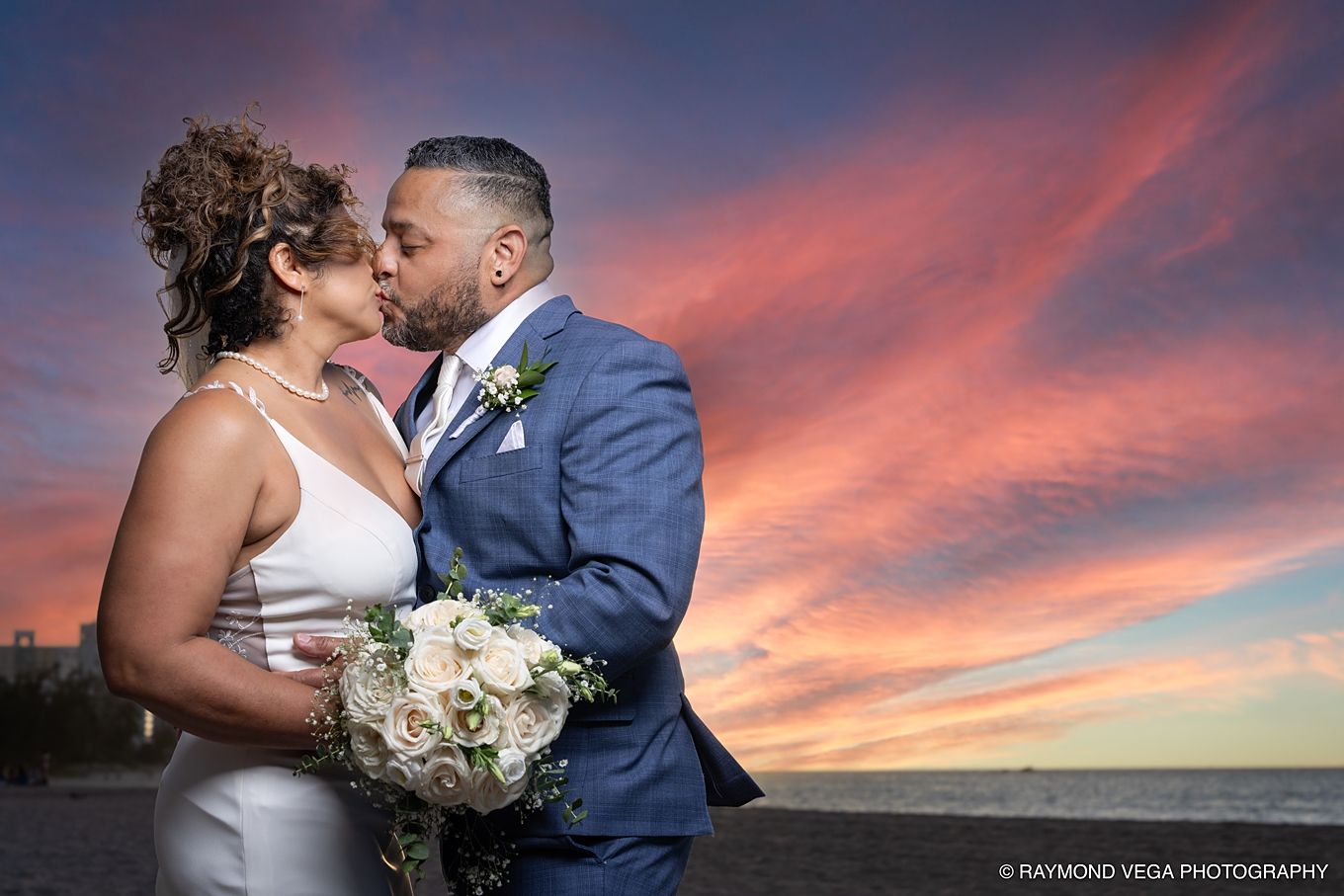 Newlywed couple sharing a romantic kiss on the beach at sunset, bride holding white bouquet, groom in blue suit. at Courtyard by Marriott