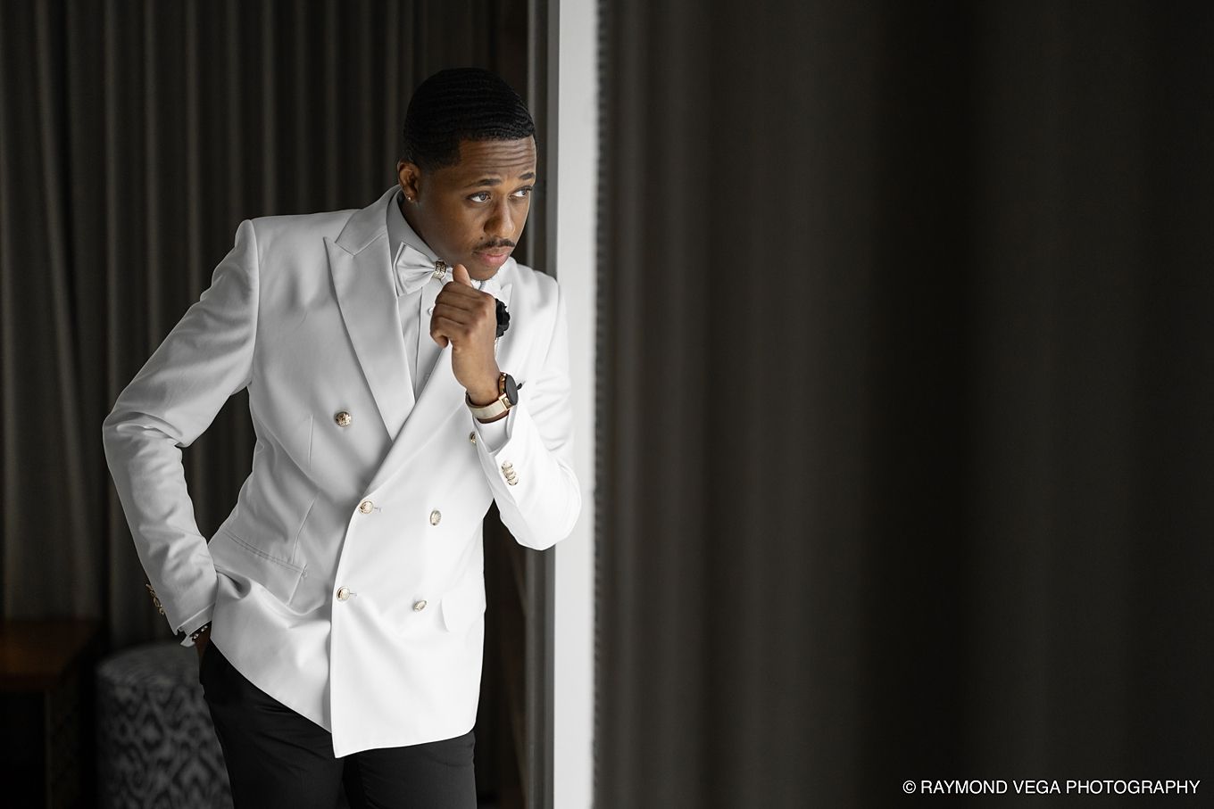 A stylish man in a white double-breasted blazer adjusts his tie near a window in a dramatic, moody portrait.