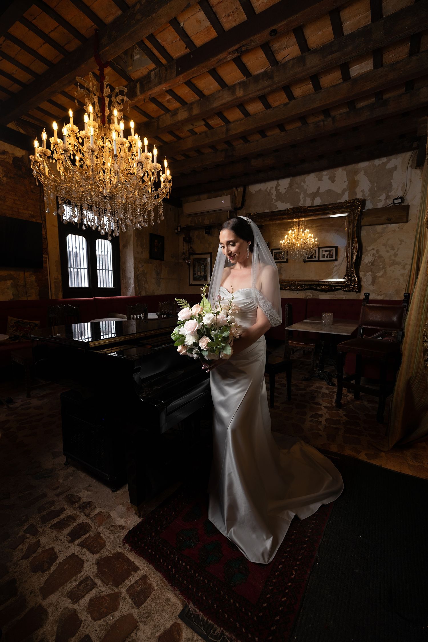 Bride in white gown and veil holds bouquet in rustic venue with stone walls and elegant chandelier lighting.