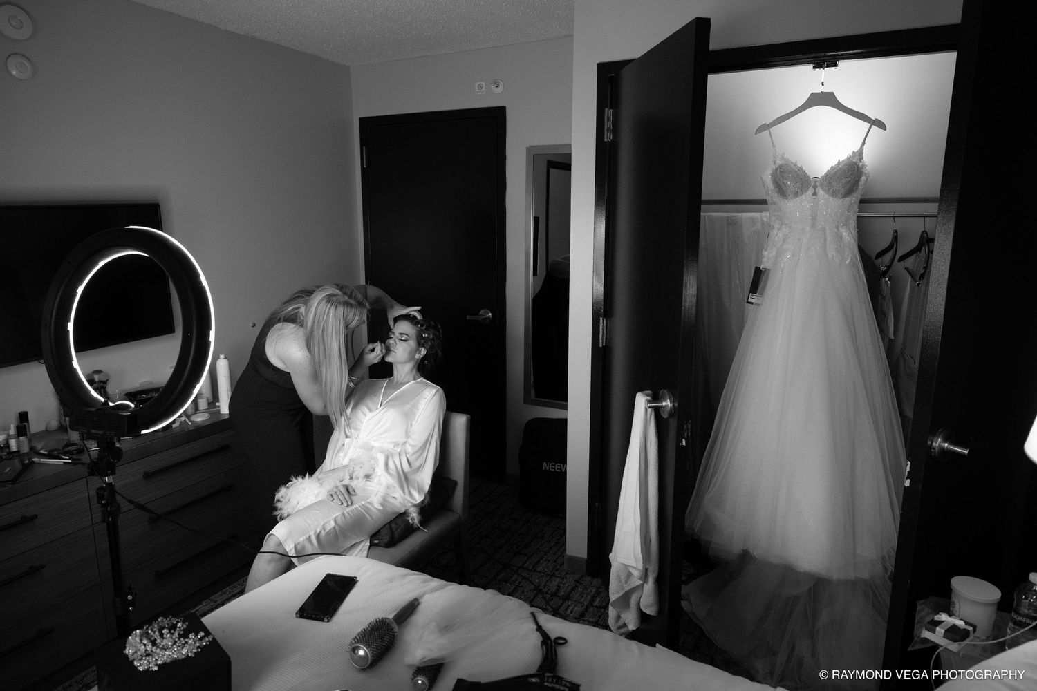 Black and white photo of a bride getting ready, with her wedding gown hanging in the background.