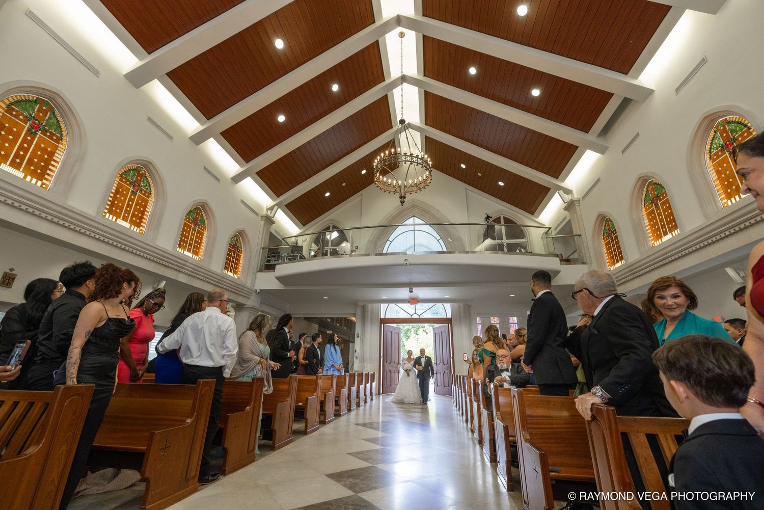 Elegant church wedding ceremony with guests standing in wooden pew aisles beneath a vaulted ceiling with stained glass windows.
