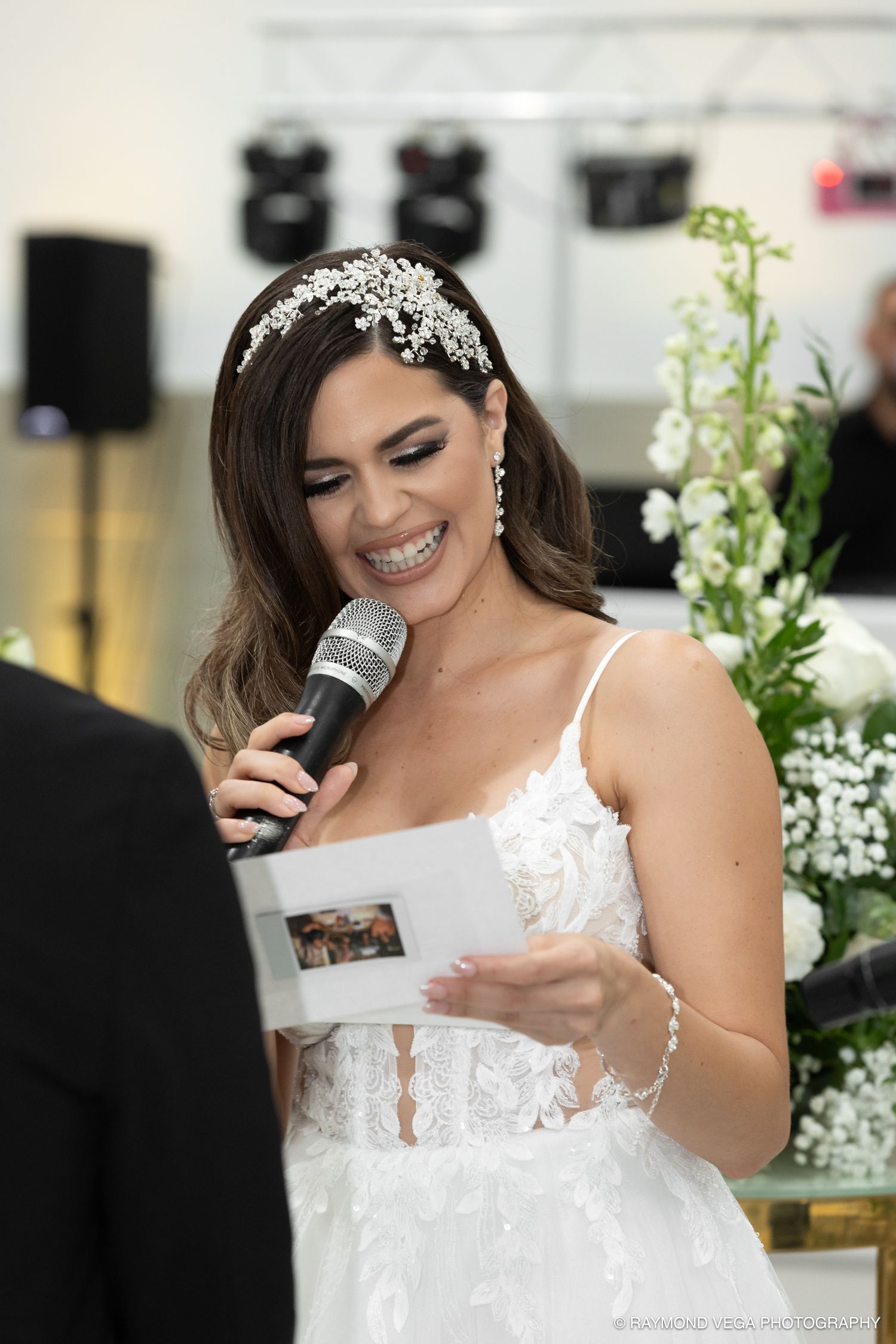 Smiling bride in white lace gown and floral headpiece reads vows into microphone at elegant wedding ceremony.