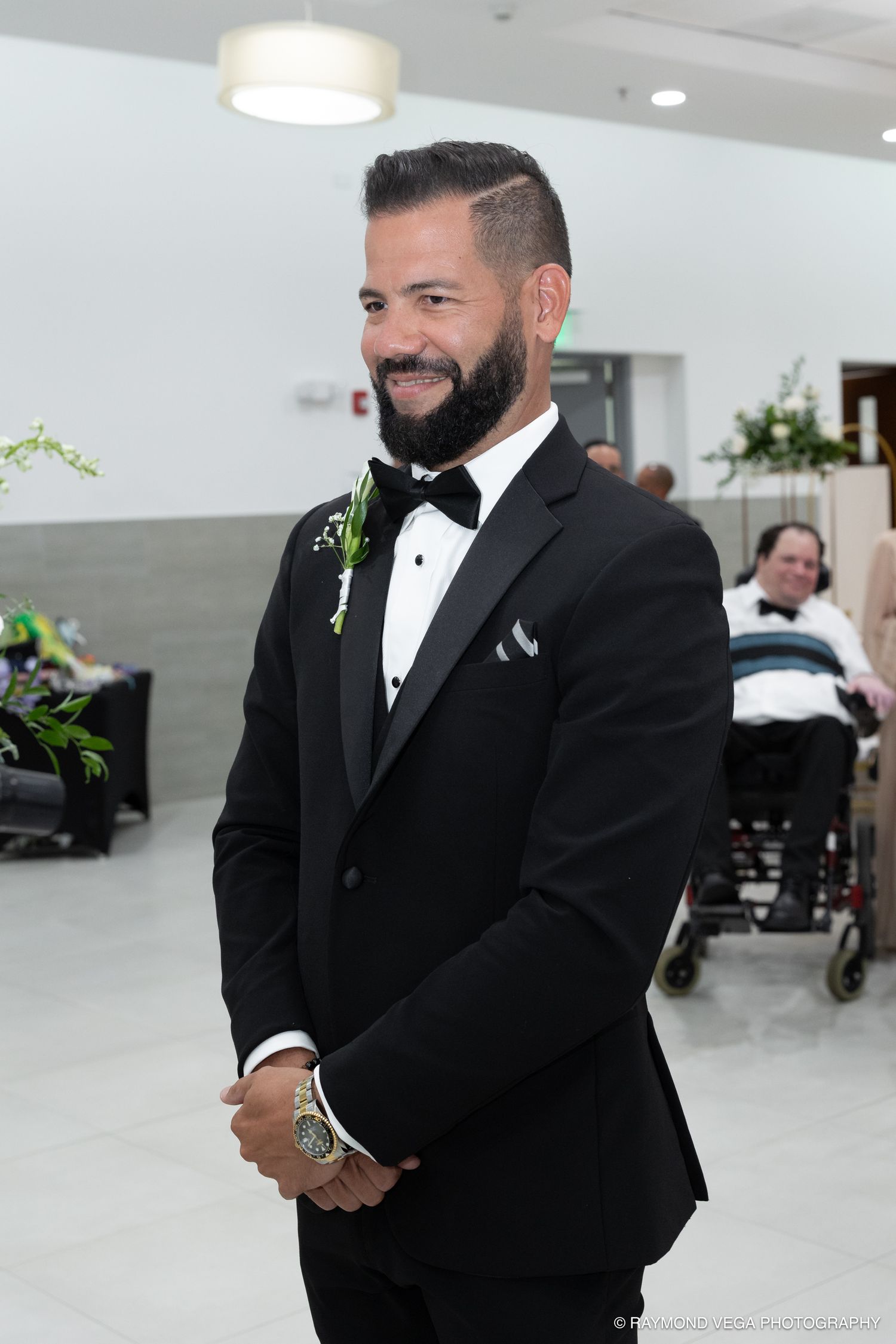 Smiling groom in black tuxedo with bow tie and boutonniere stands at wedding venue.