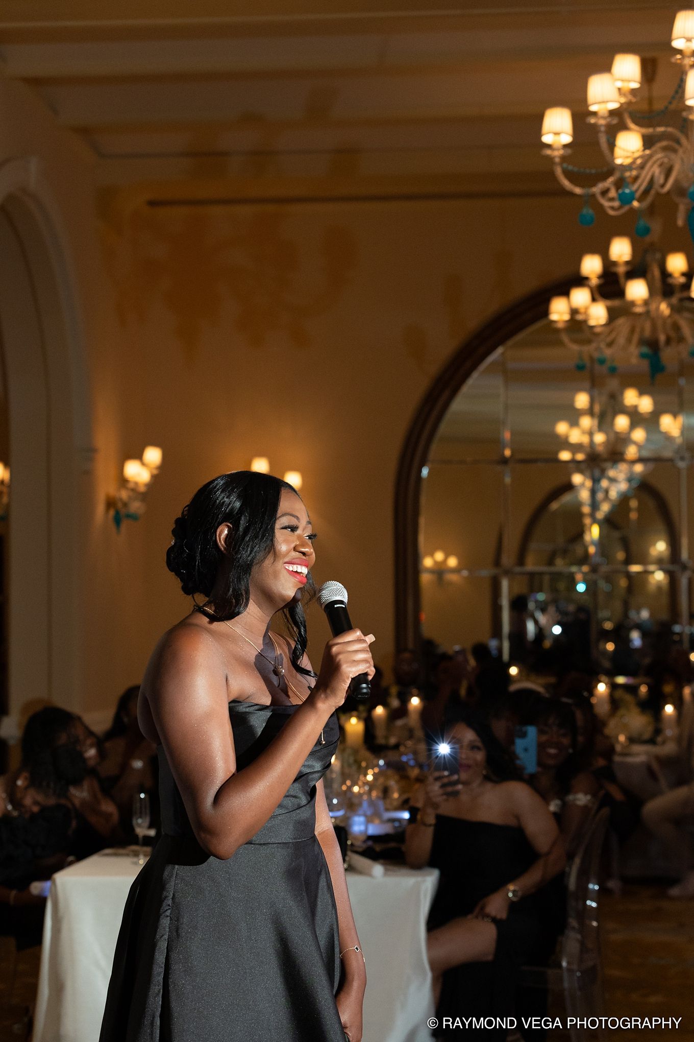 A woman in a gray gown smiles while giving a speech with a microphone at an elegant candlelit reception hall.
