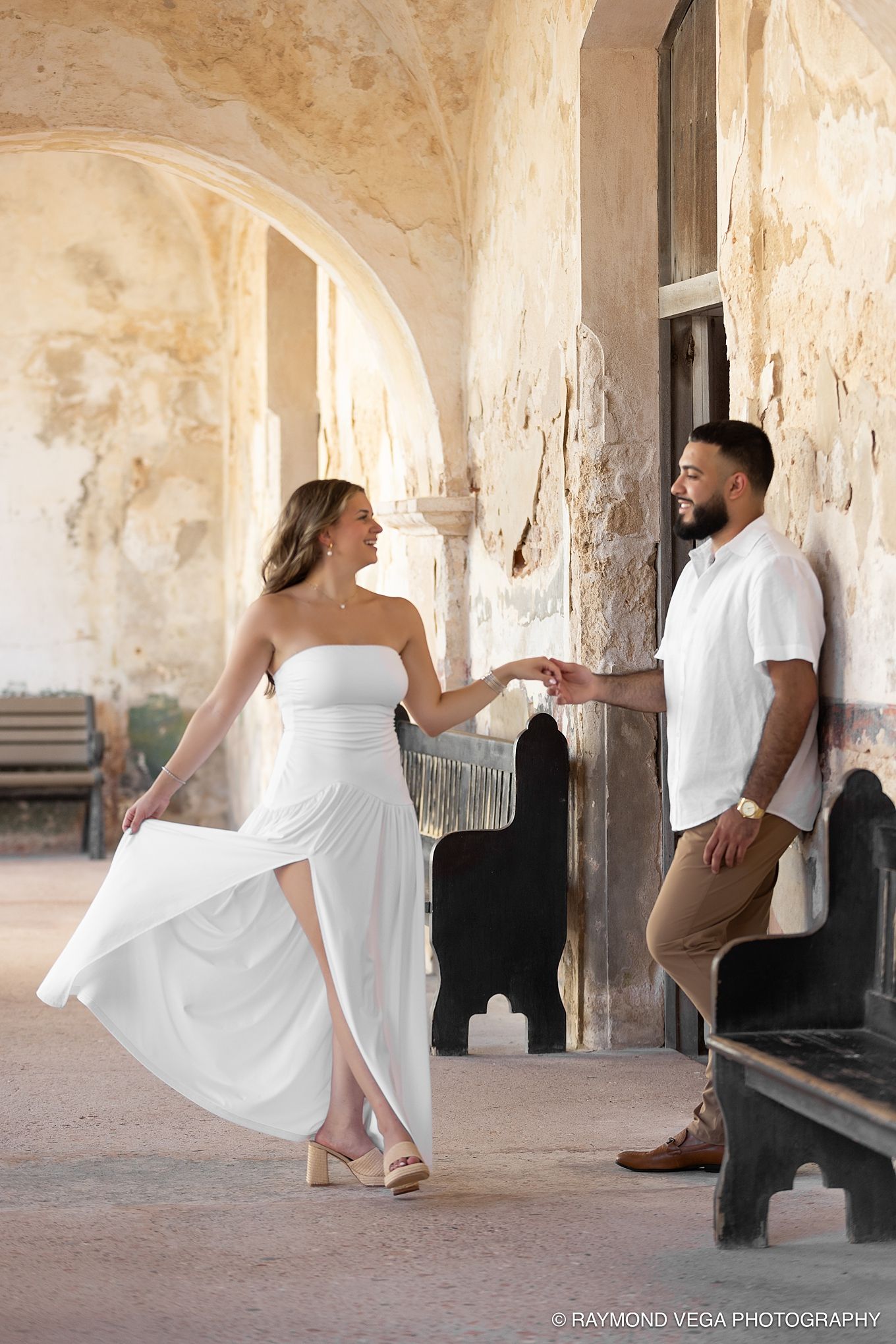 Couple in white outfits holding hands at a historic stone archway, captured by Raymond Vega Photography. Castillo de San Cristóbal