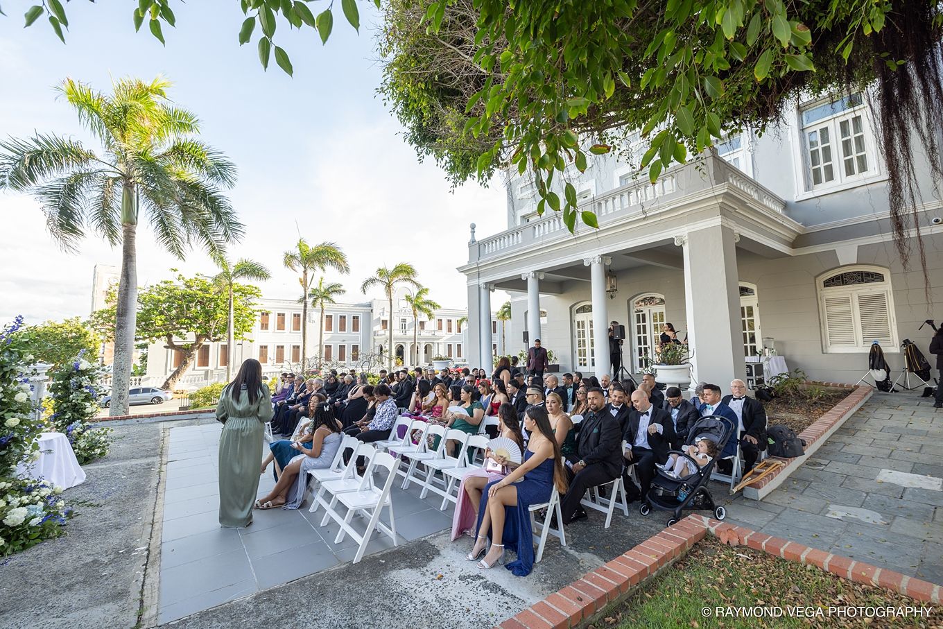 Outdoor wedding ceremony with guests seated in white chairs on a patio beside an elegant colonial building surrounded by palm trees.