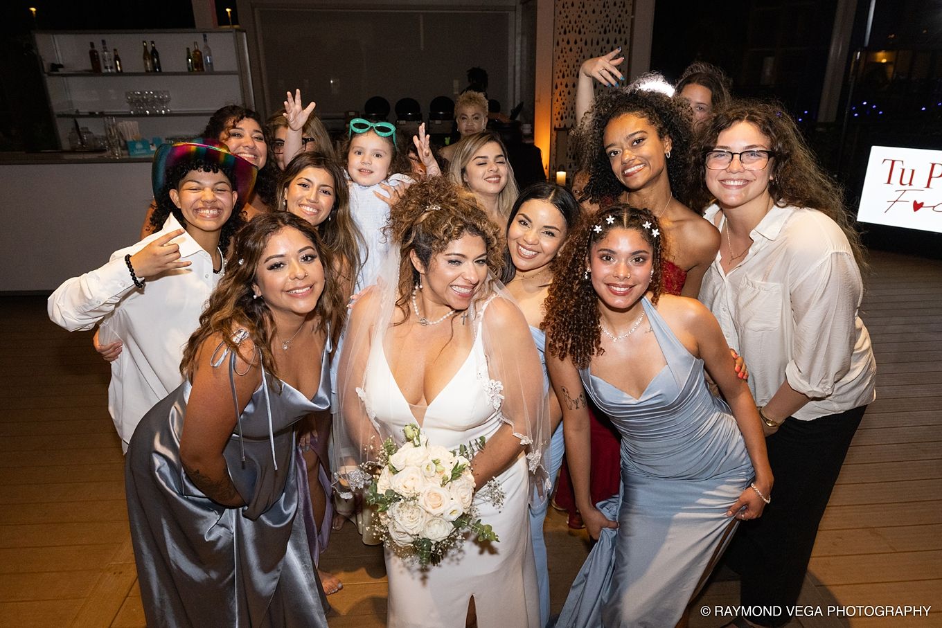 Bride in white gown poses with bridesmaids and friends at wedding reception, smiling and celebrating together.
