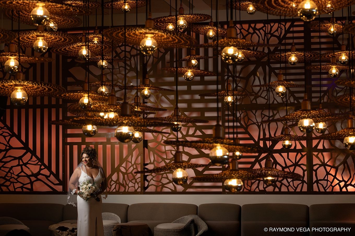 Bride in white gown holding bouquet stands before ornate metalwork wall with warm glowing pendant lights.