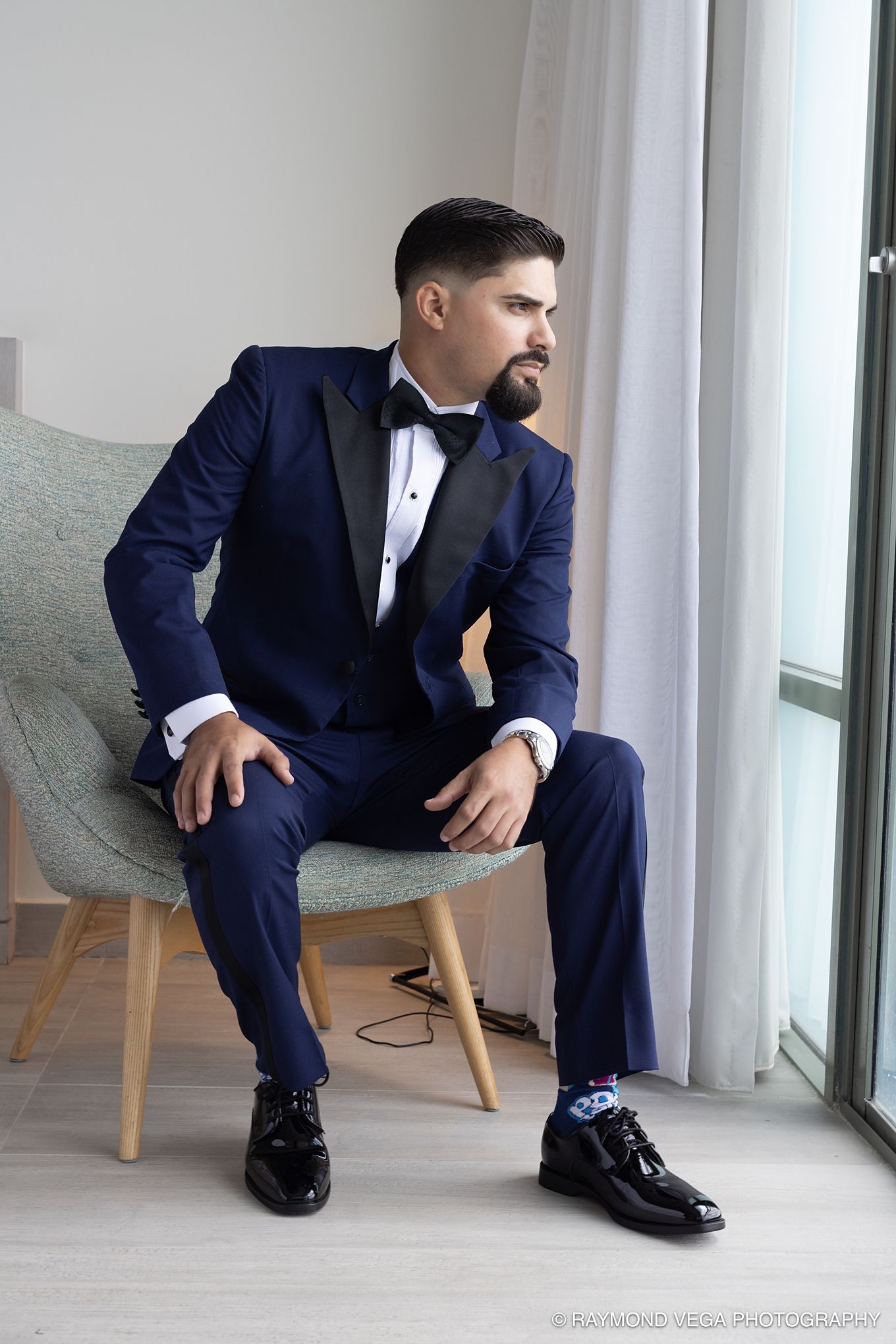 Man in navy blue tuxedo with bow tie sitting elegantly in a chair by a window, wearing patent leather shoes. Hotel la Concha Wedding