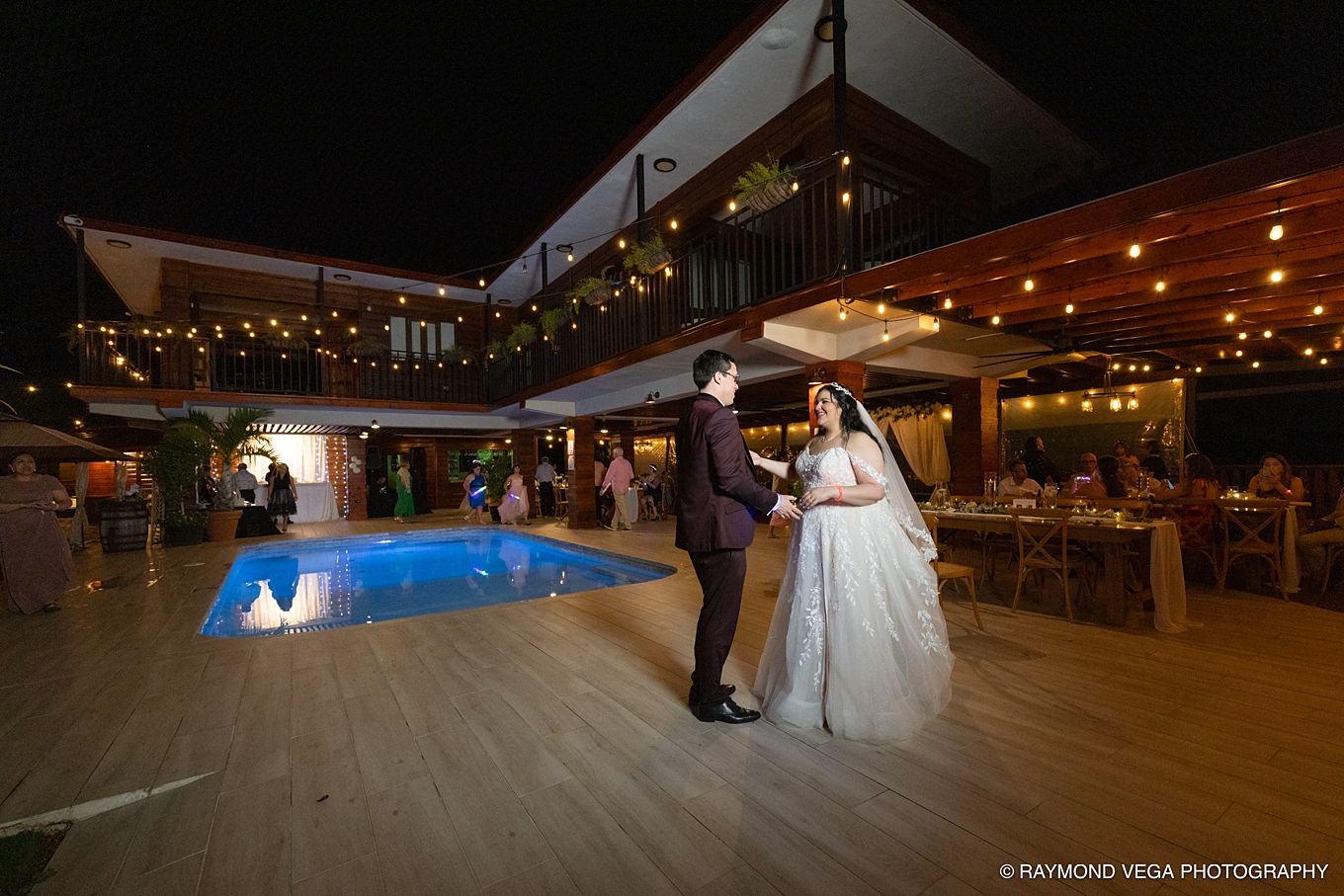 Bride and groom share first dance beside glowing pool at elegant outdoor wedding reception with string lights at night.
