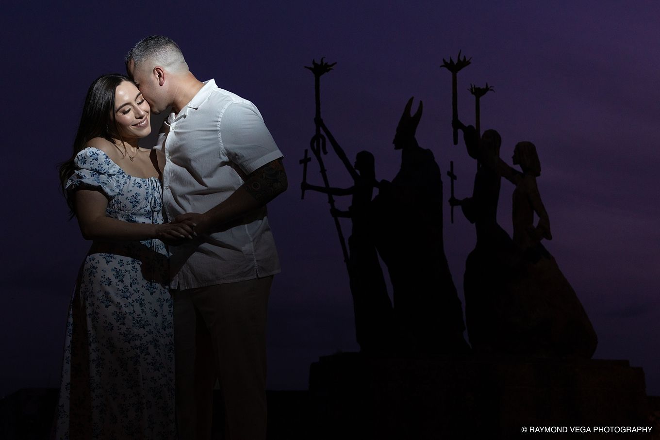 Romantic couple embracing at dusk with dramatic silhouette sculpture backdrop in purple twilight sky. - Love Story