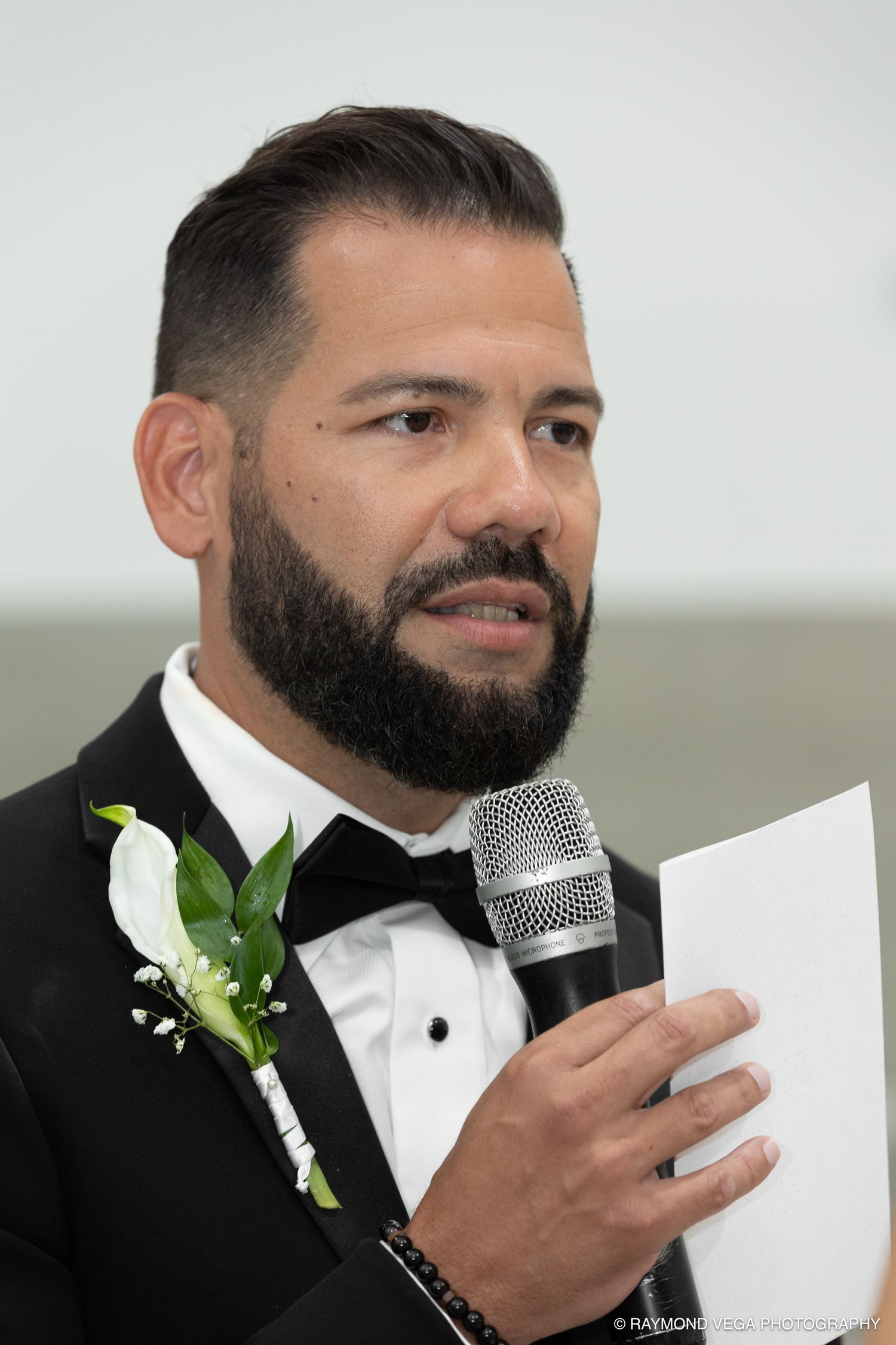 A bearded groom in a tuxedo with a white boutonniere speaks into a microphone while holding a paper at a wedding.