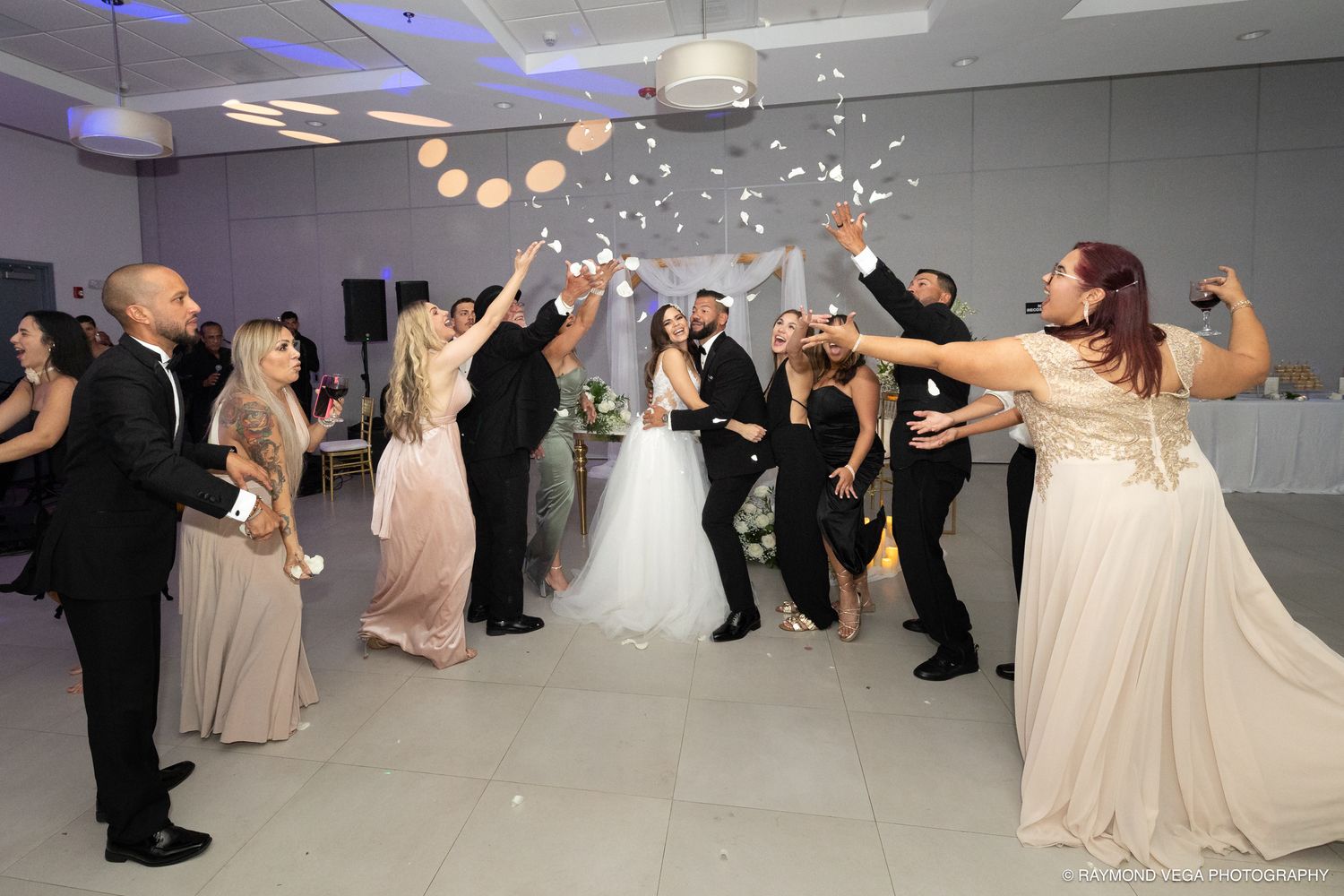 Wedding guests tossing confetti over bride and groom in elegant reception hall with soft pink lighting.