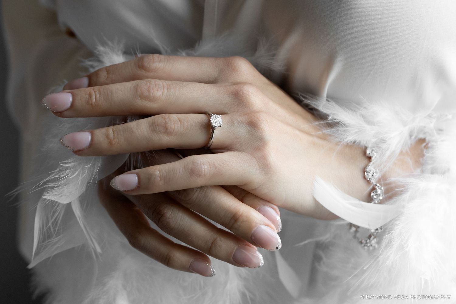 Close-up of elegant hands wearing a diamond engagement ring and bracelet, with soft white feathers in background.