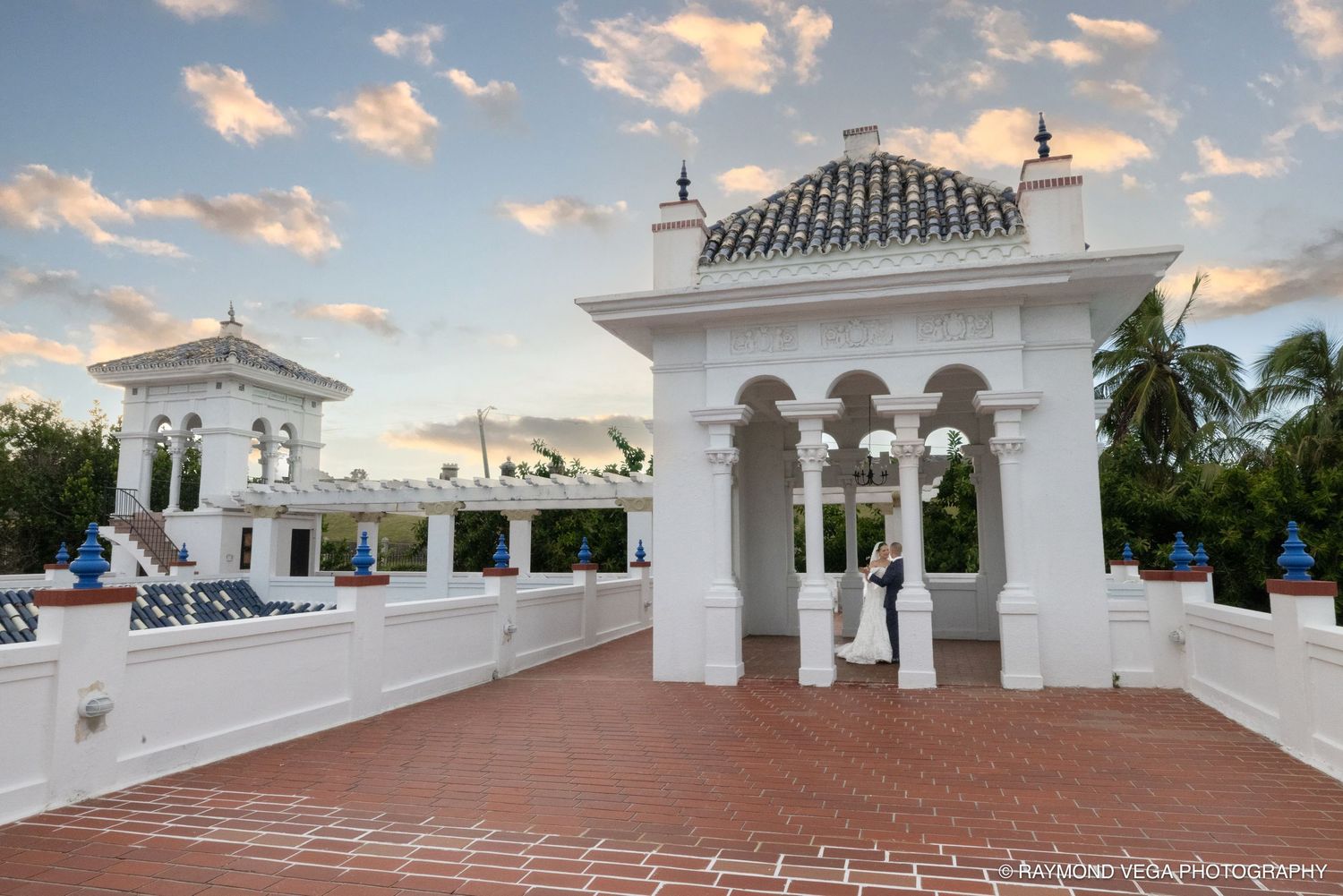 Elegant white gazebo on a brick terrace at a tropical wedding venue with palm trees and a colorful sunset sky.