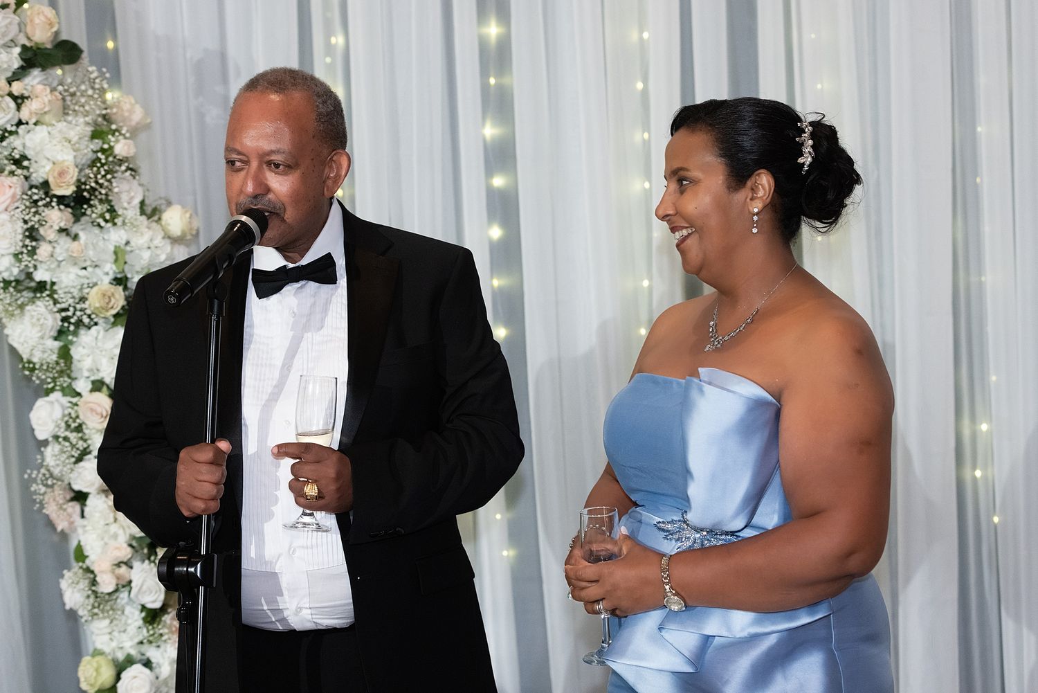 Man in tuxedo speaking at microphone beside woman in blue strapless gown at elegant wedding reception.
