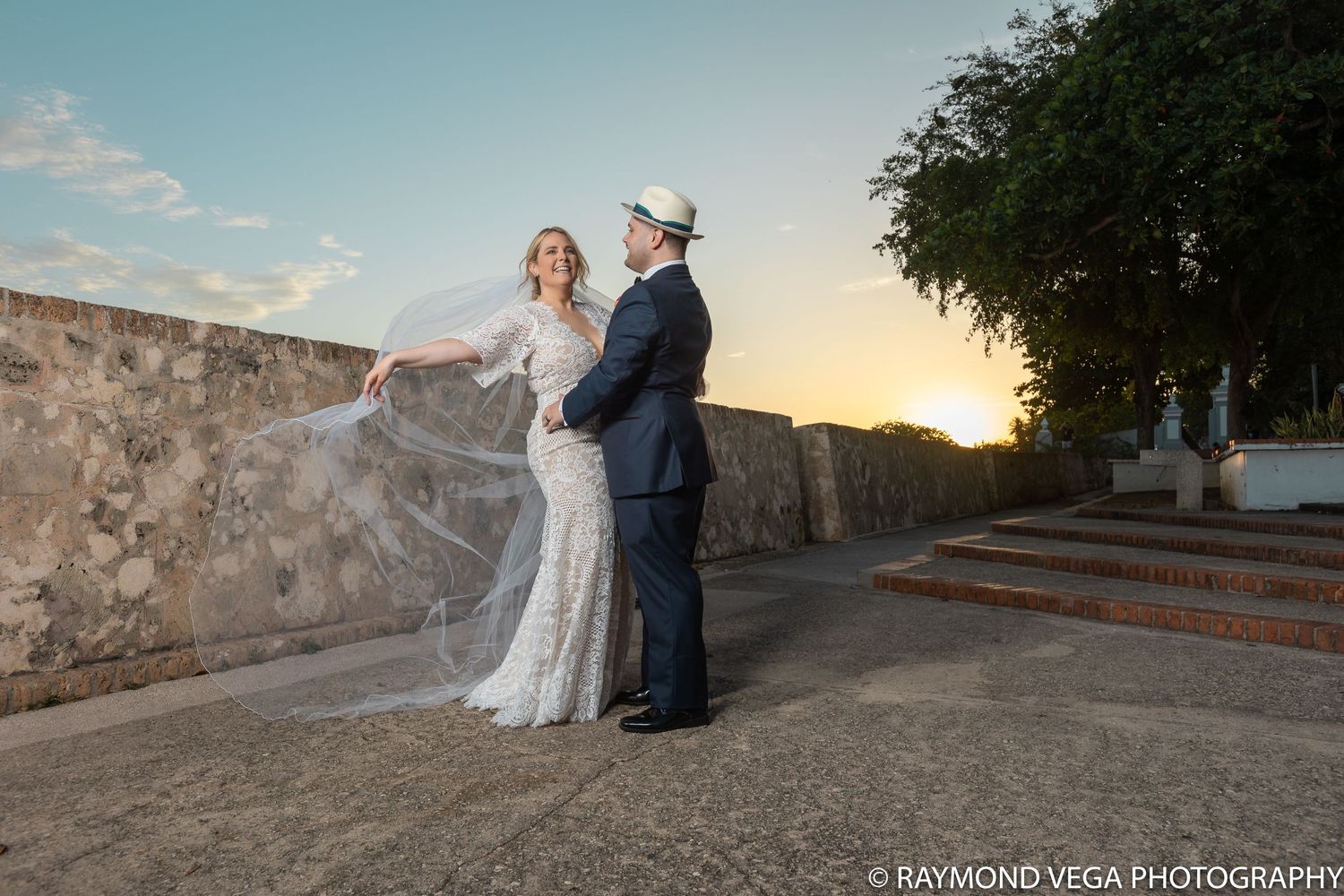 Bride and groom share romantic moment at sunset near stone wall, her veil flowing in the breeze.