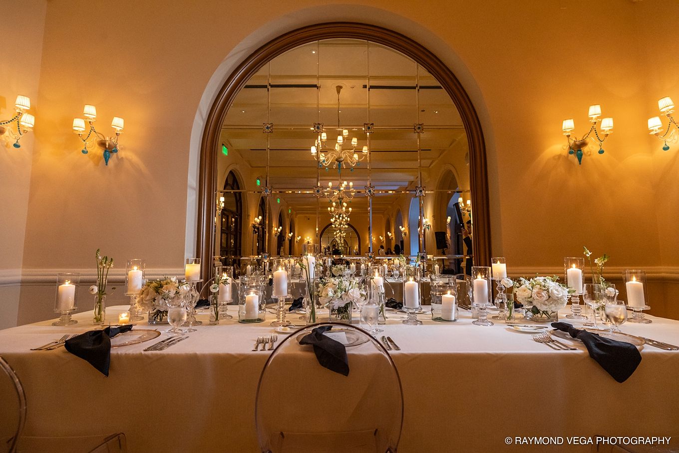 Elegant wedding reception hall with long white table settings, black napkins, candles, and a grand mirror reflecting a chandelier.