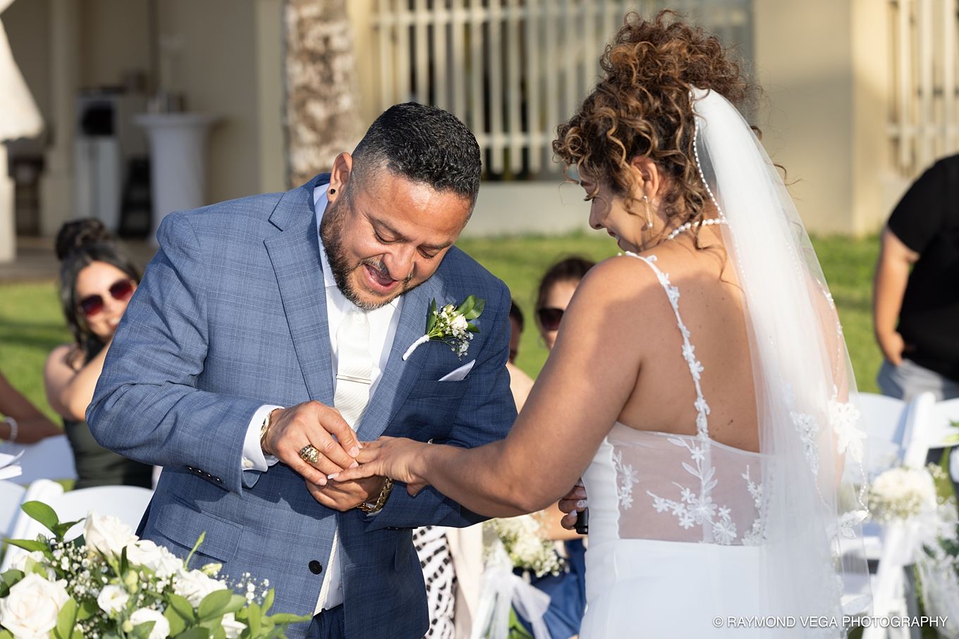 Groom in blue suit placing wedding ring on bride's finger during outdoor ceremony with white floral arrangements. at Courtyard by Marriott