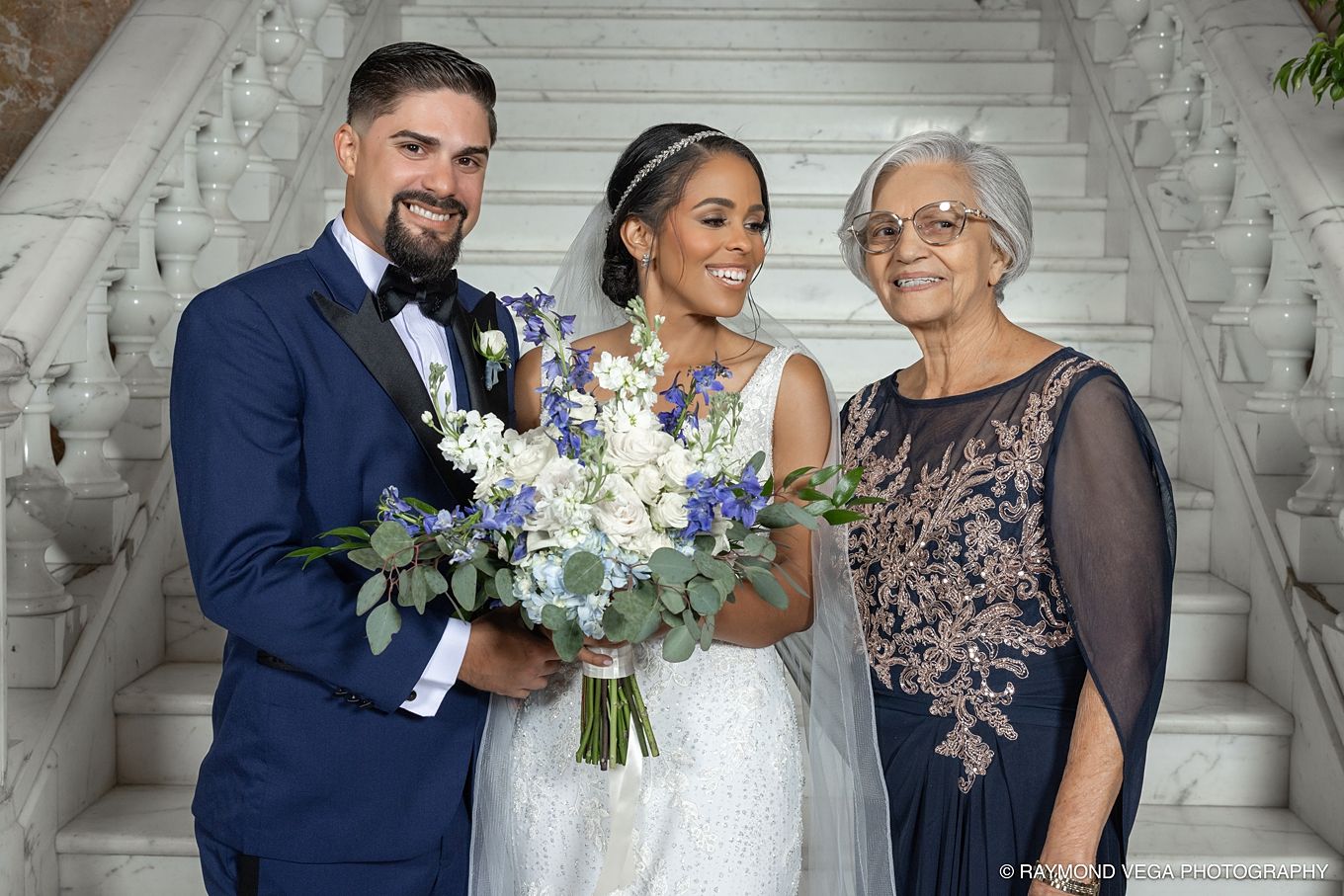 Bride and groom pose with elderly woman on marble staircase, bride holding blue and white floral bouquet. Antiguo Casino Puerto Rico