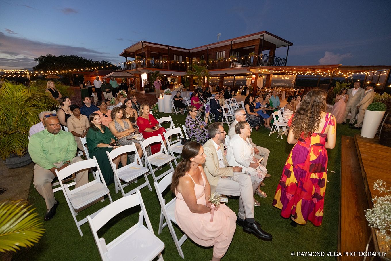 Guests seated in white folding chairs at an outdoor evening wedding ceremony on a rooftop venue with string lights.