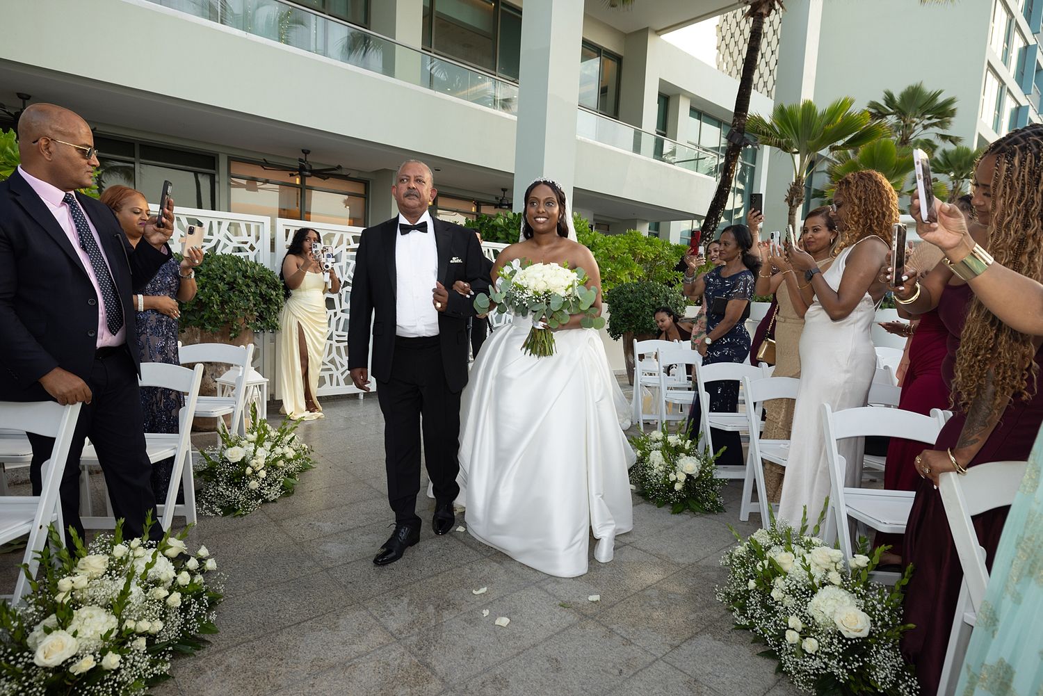 Bride and groom walk down outdoor wedding aisle lined with white floral arrangements and cheering guests.