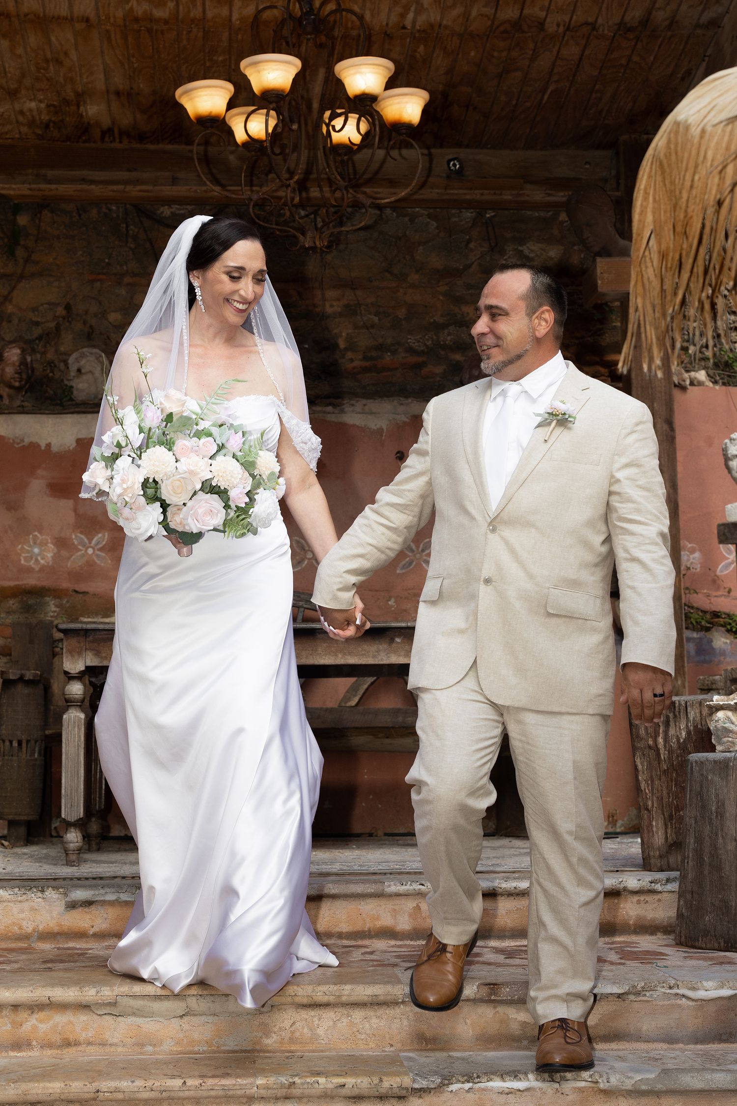 Bride in white gown holding bouquet walks hand-in-hand with groom in beige suit down stone steps at wedding venue.