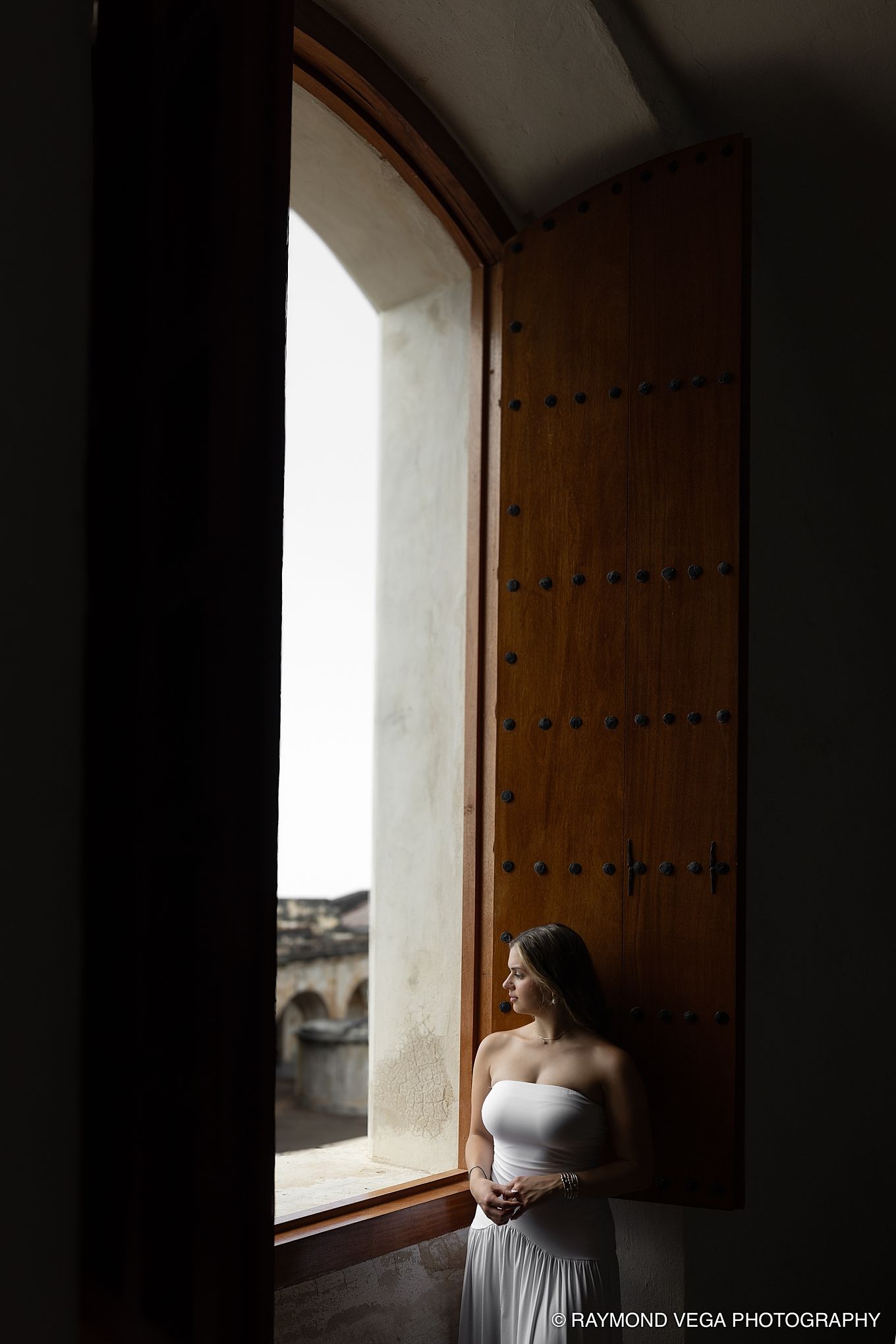 Bride in white strapless gown stands by ornate wooden door with arched window in dramatic light.
