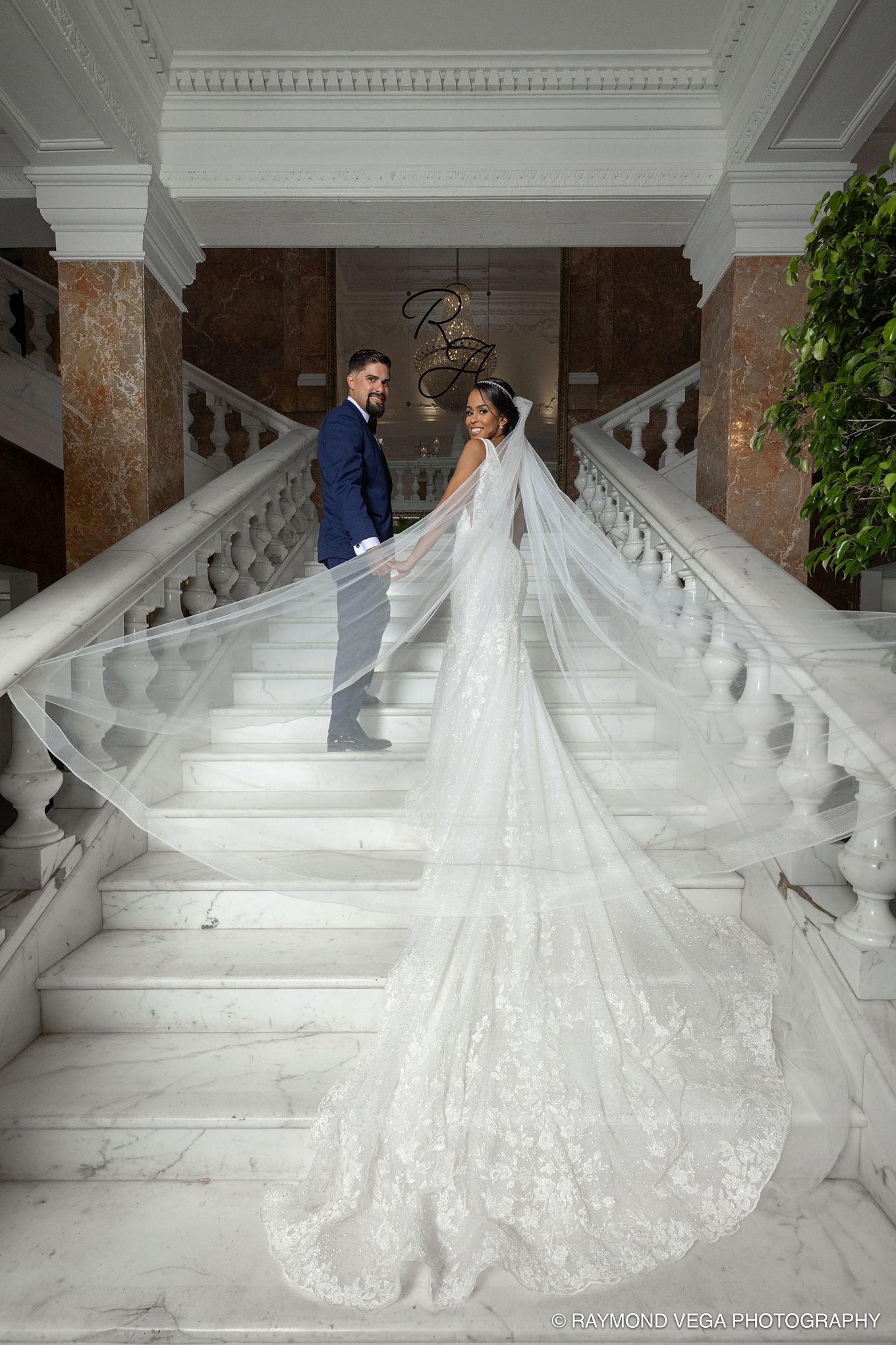 Bride and groom holding hands on grand marble staircase, bride wearing stunning lace gown with cathedral veil flowing down steps.