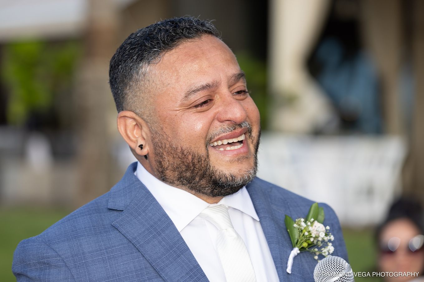 Smiling groom in blue suit with boutonniere laughing joyfully at outdoor wedding ceremony.