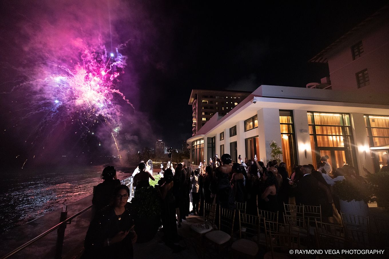 Crowd watches vibrant purple fireworks burst over a waterfront venue at night during an outdoor celebration event.