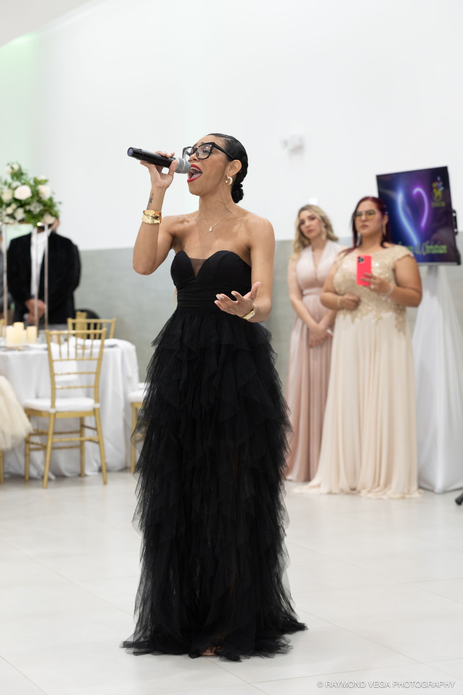 A woman in a black strapless tulle gown singing into a microphone at an elegant wedding reception.