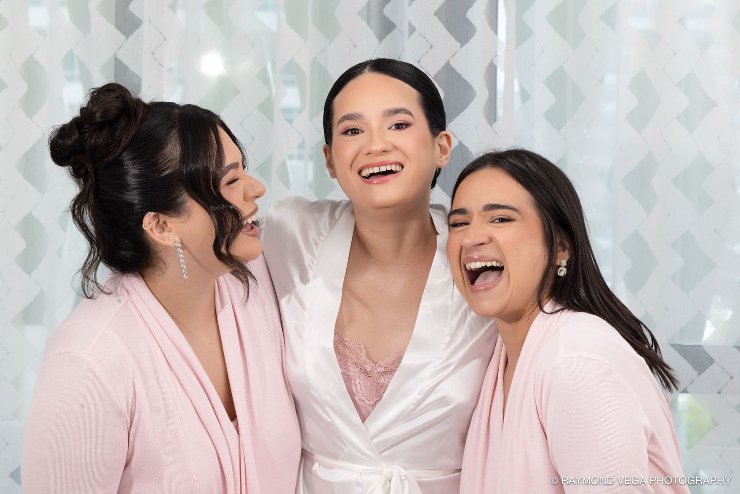 Three women in pink robes laugh joyfully together, with the bride in a white satin robe at the center.