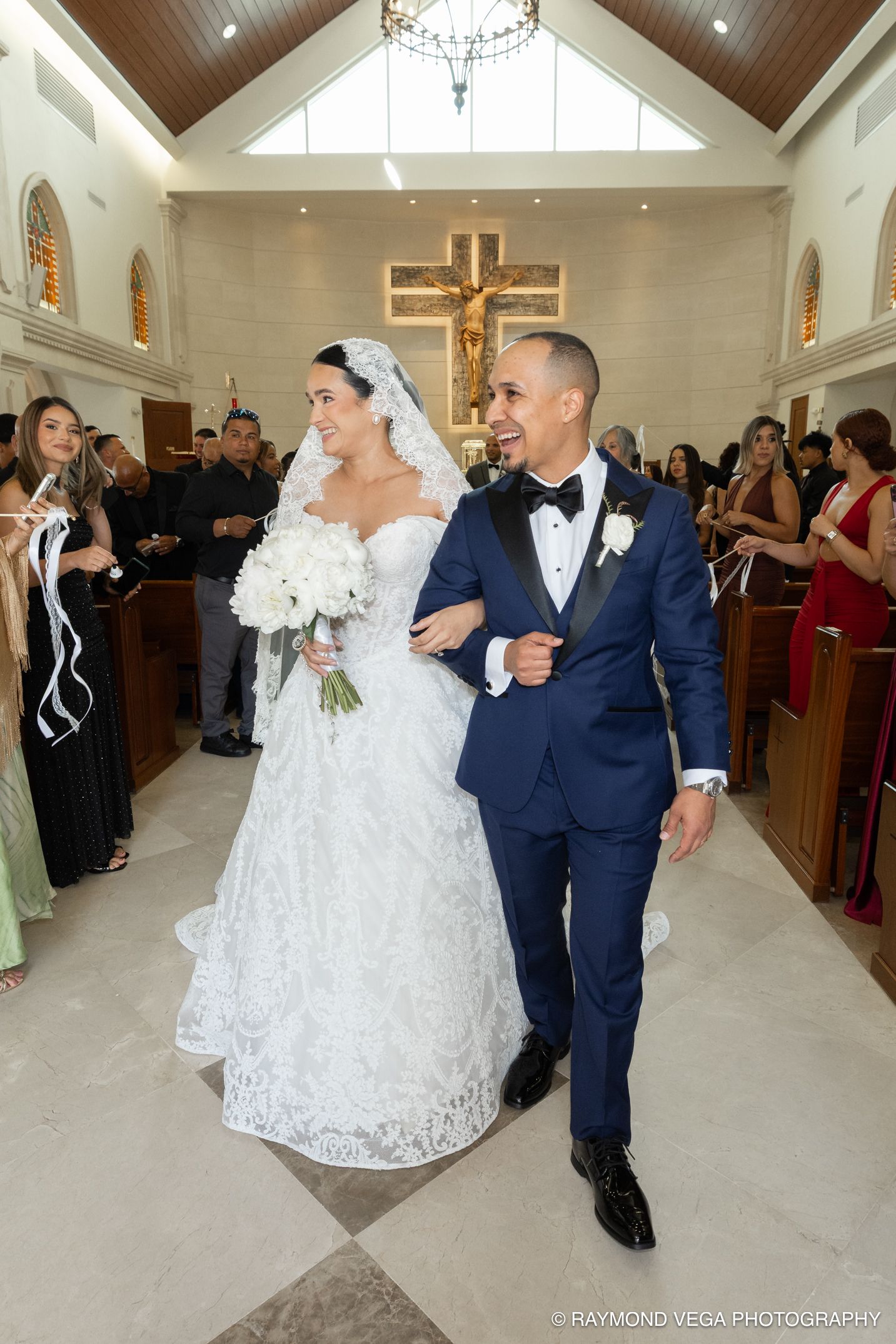 Bride and groom walk down church aisle after wedding ceremony, bride in lace gown holding white bouquet, groom in navy tuxedo.