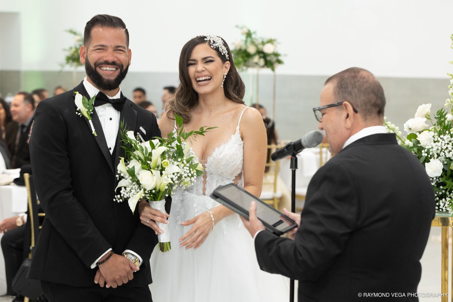 Happy couple smiling during wedding ceremony with officiant, bride holding white floral bouquet in elegant venue.