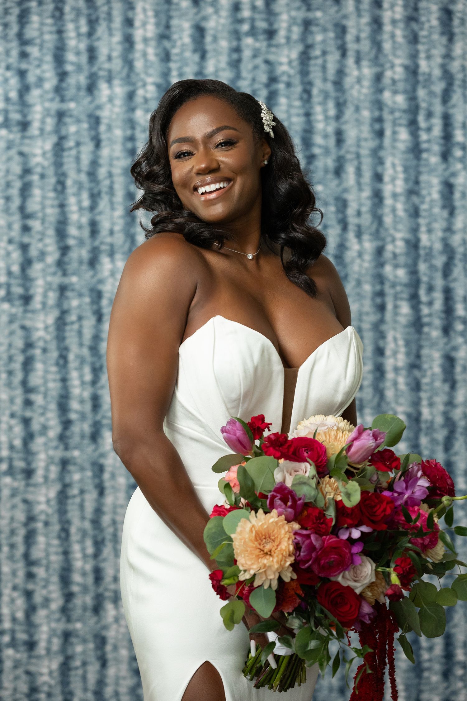 Smiling bride in white strapless gown holding vibrant red and pink bouquet against blue backdrop.
