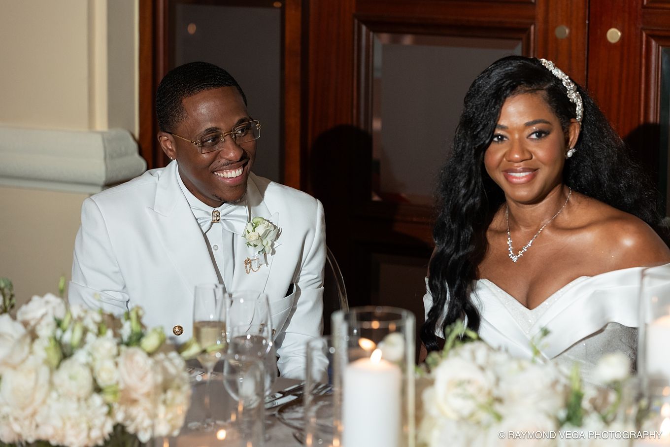 Smiling bride and groom in white attire seated at wedding reception table with floral arrangements and candles.