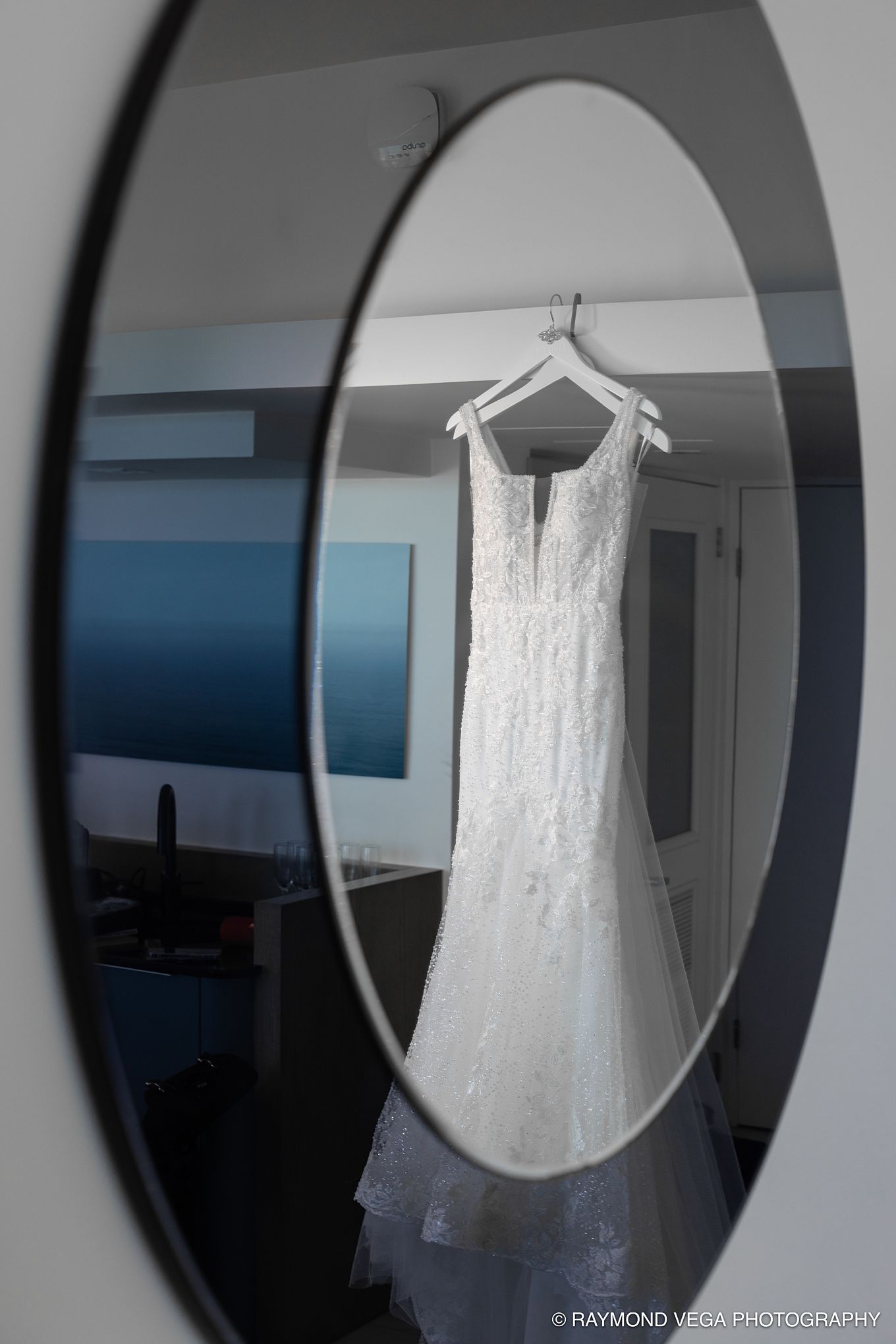A white lace wedding dress hanging on a hanger, reflected in a round black-framed mirror in a bright room.