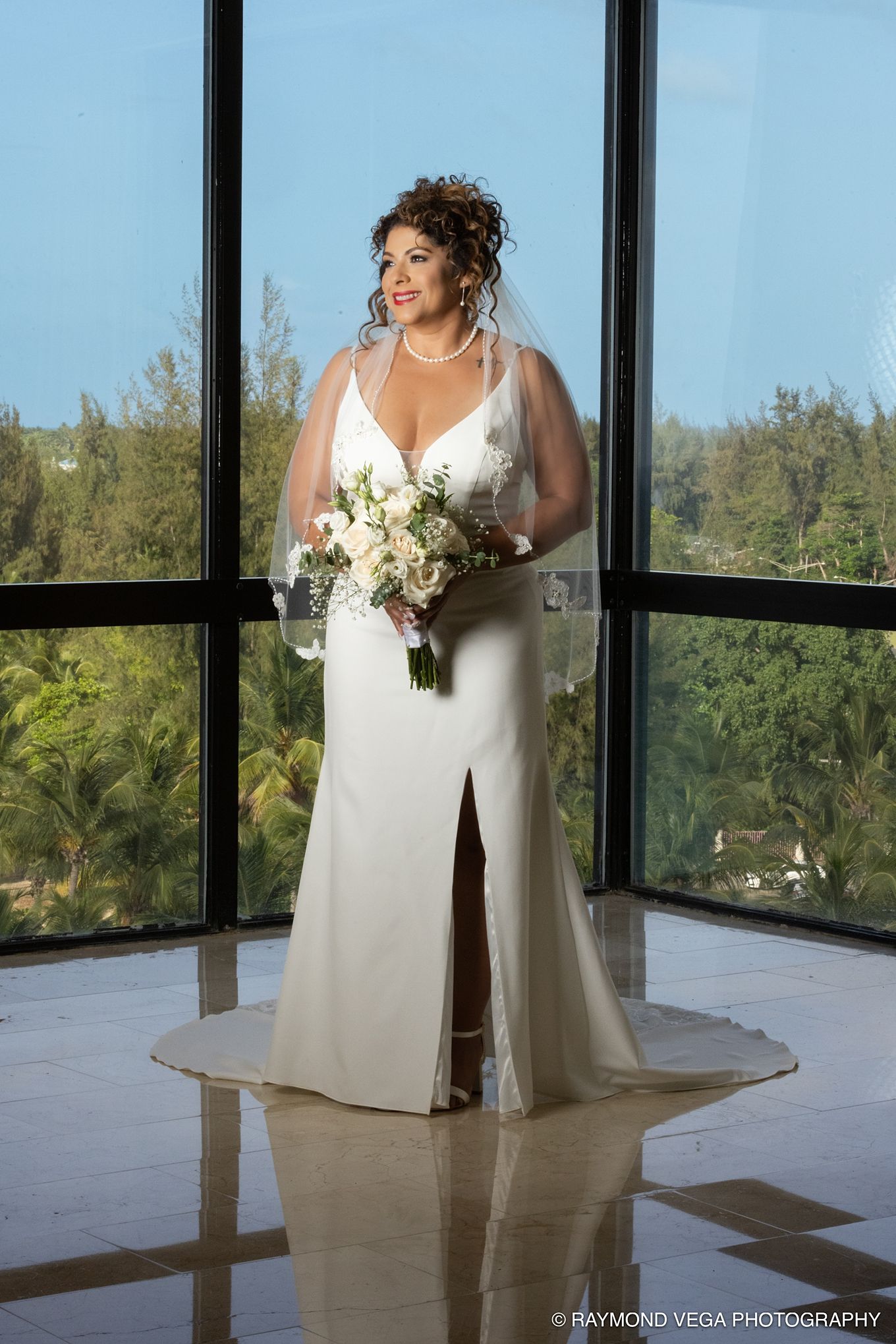 Bride in white gown with veil holds floral bouquet, standing before large windows with tropical view.
